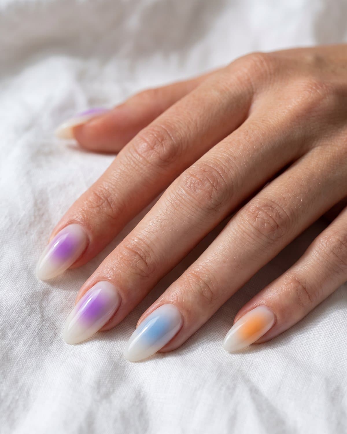 Almond aura nails with soft airbrushed glows of purple, blue and sunset orange in the center of a milky base, on white linen
