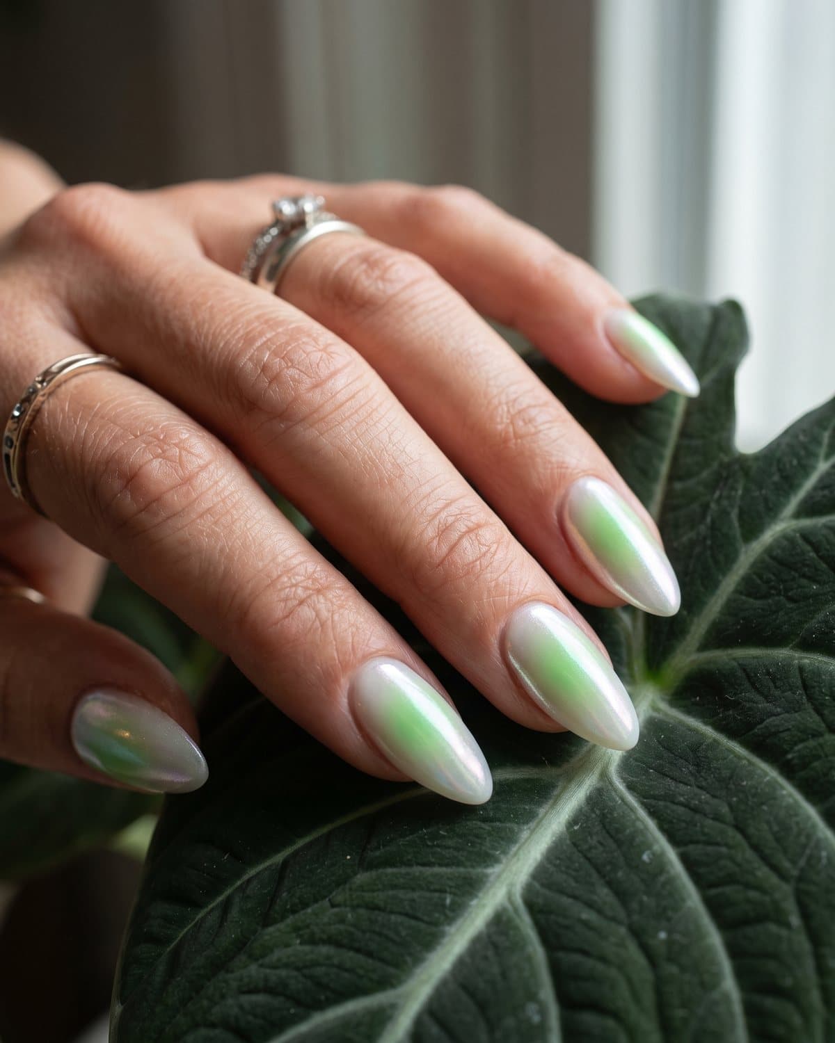 Almond nails with a green aura glow on a milky base and aura nails topped with iridescent pearl chrome, beside a green leaf