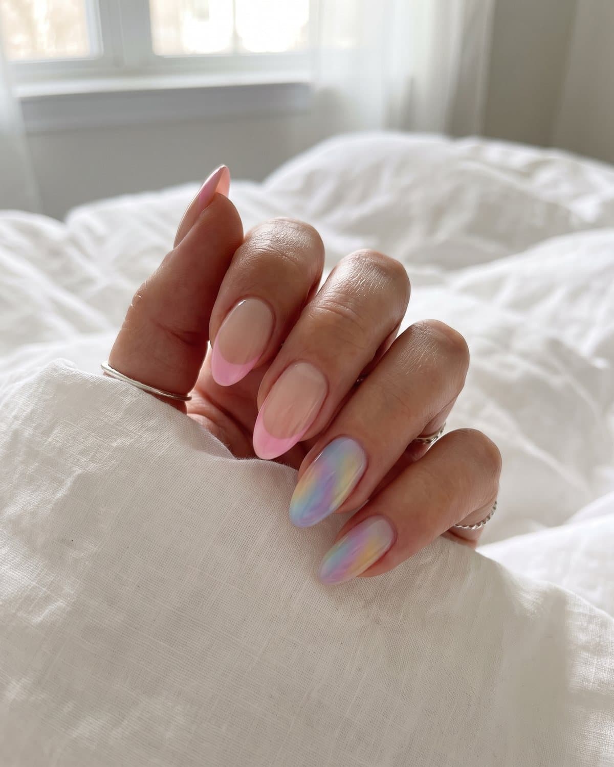 Almond nails with a sheer nude base and pink aura French tips mixed with mismatched rainbow aura nails, on white bedding