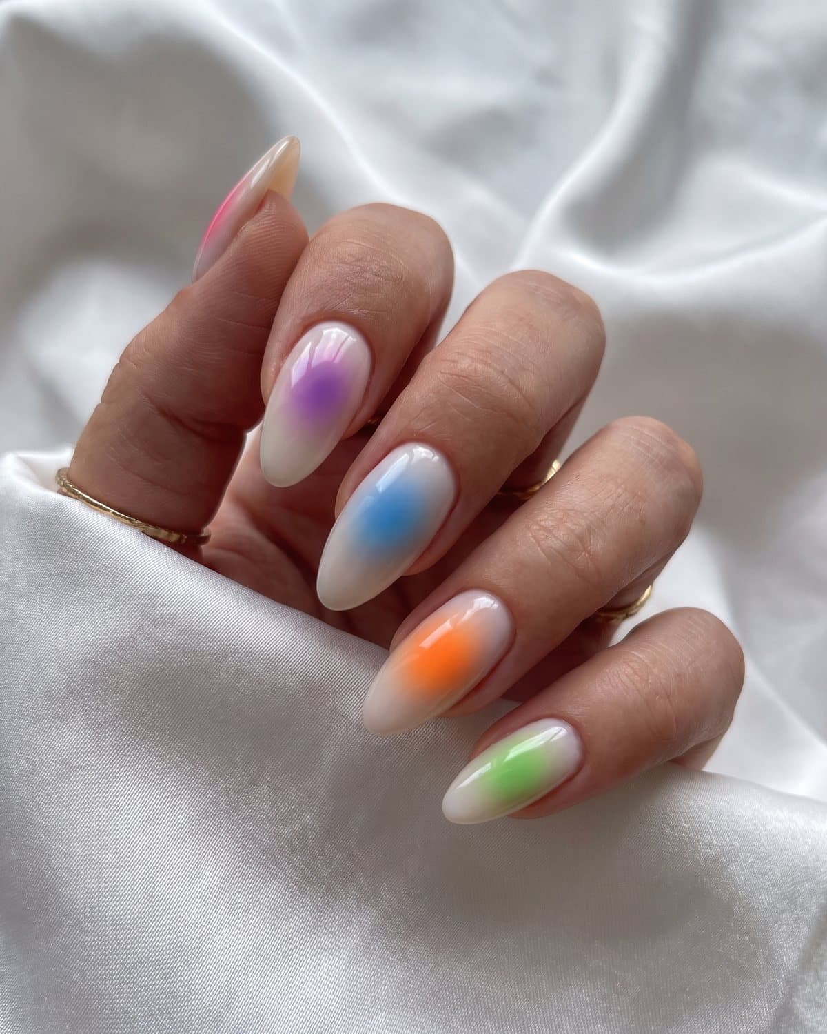 Almond aura nails with soft airbrushed glows of pink, purple, blue, orange and green on a milky base, on white satin