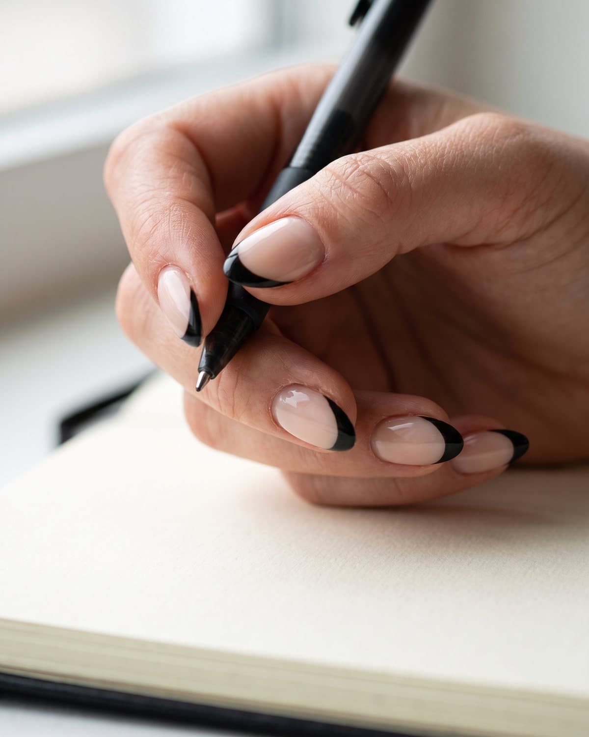 Glossy almond nails with a sheer nude base and classic black French tips, holding a black pen over a notebook