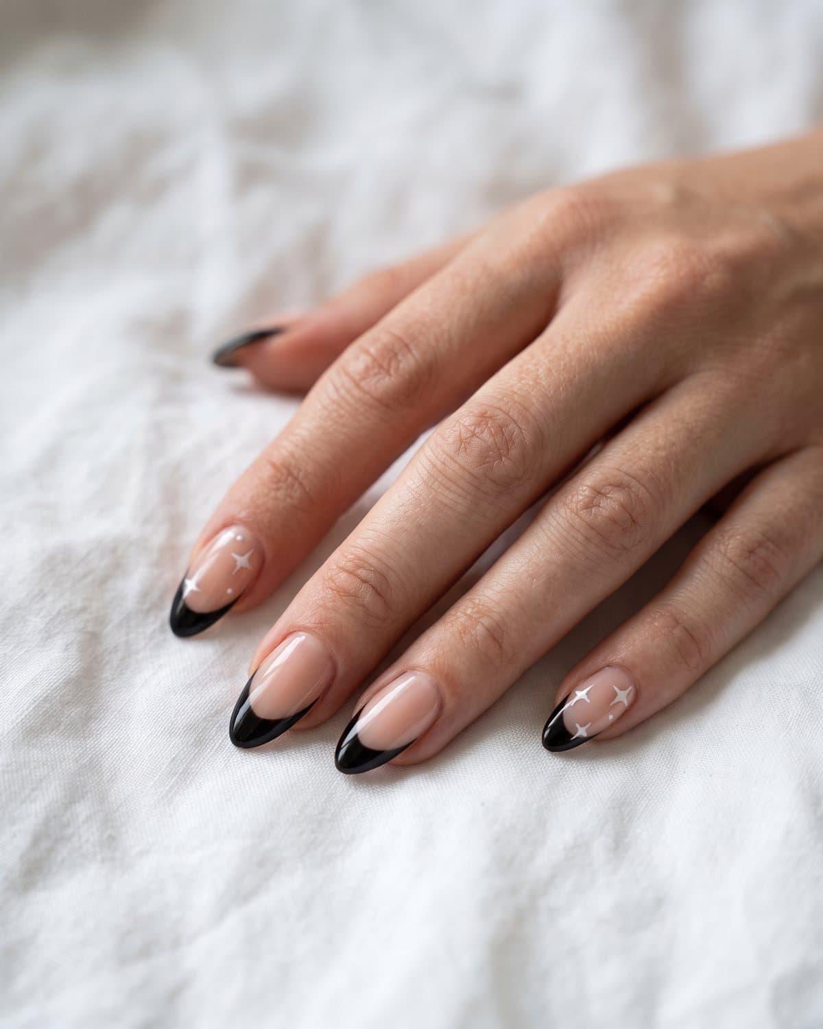 Almond nails with diagonal side-swept black French tips and black tips with tiny white stars, on white linen