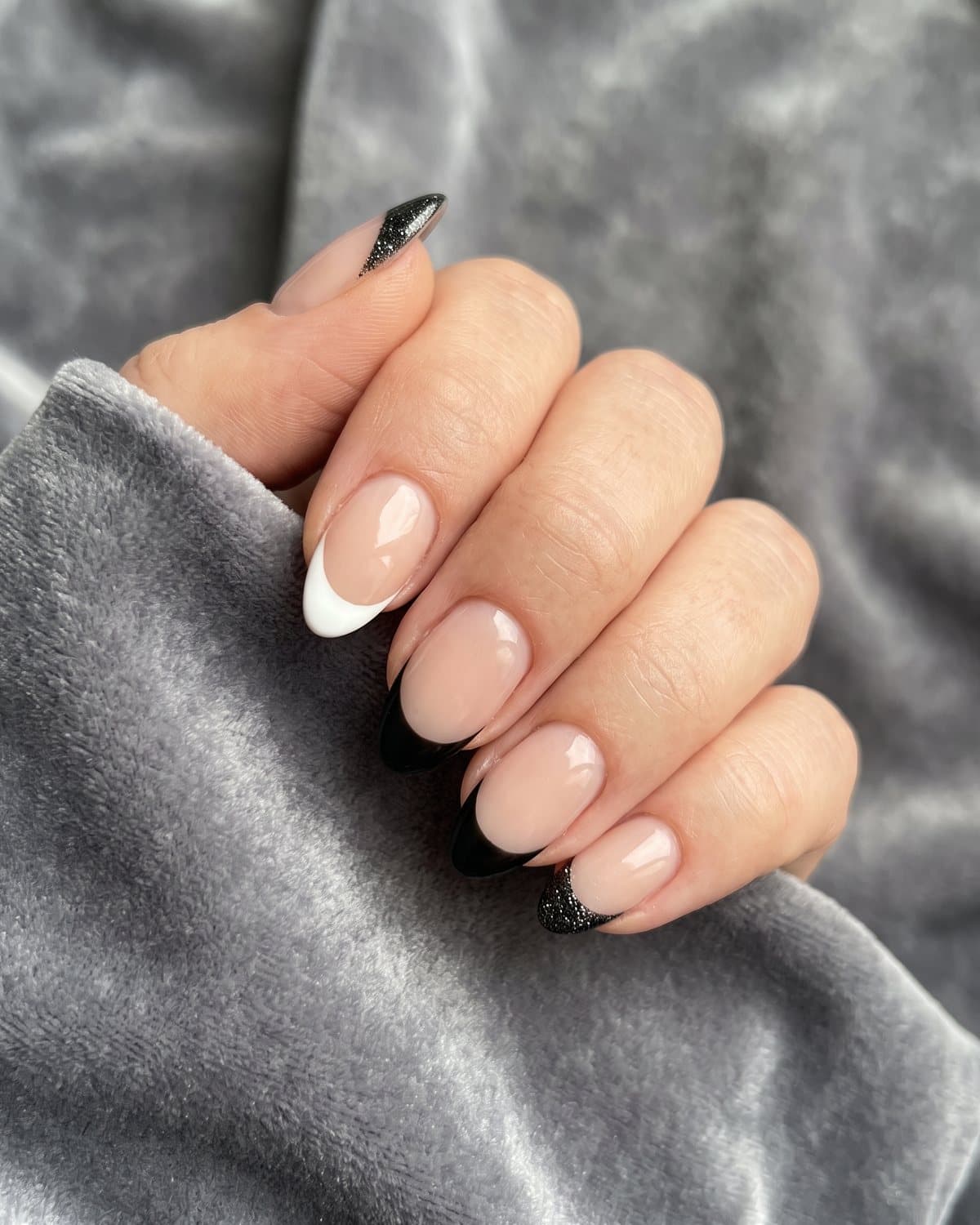 Almond nails with sparkly black glitter French tips alternating with mismatched black and white French tips, on grey velvet