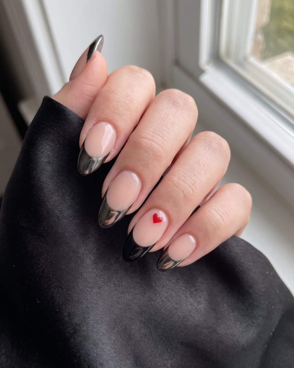 Almond nails with metallic black chrome French tips and one black tip with a tiny red heart, on black satin