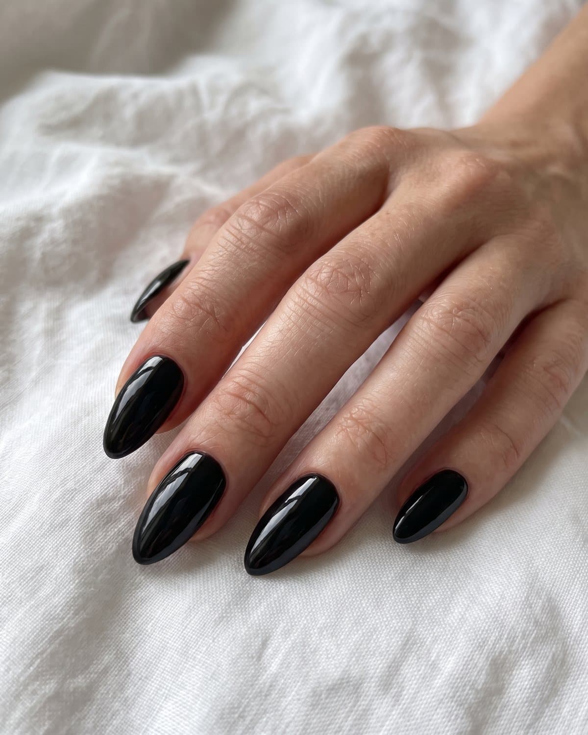 Long almond nails in a flawless high-shine glossy black, resting on white linen