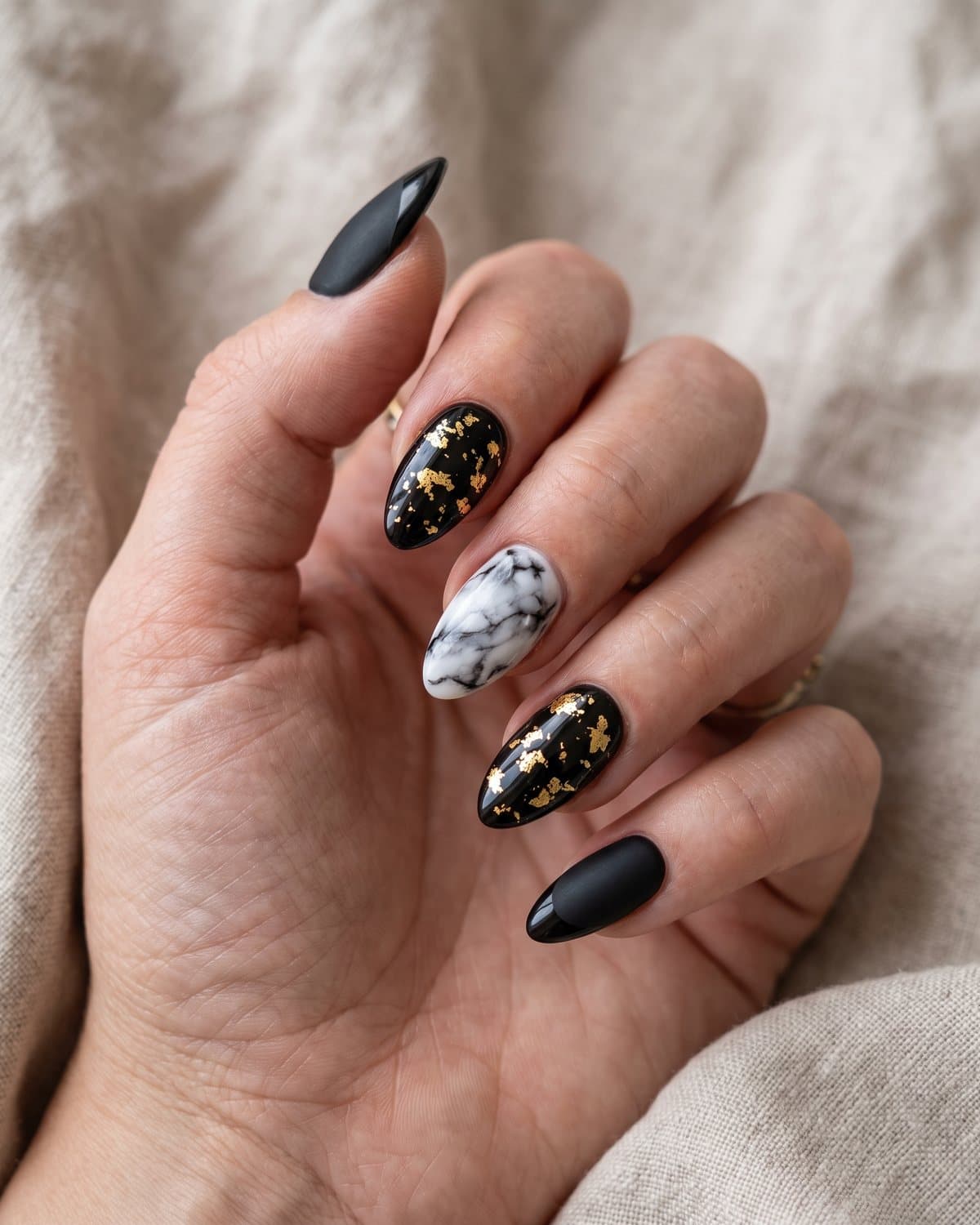 Almond nails in glossy black with gold foil flakes, black and white marble with soft veins and matte black with glossy black French tips, on beige linen