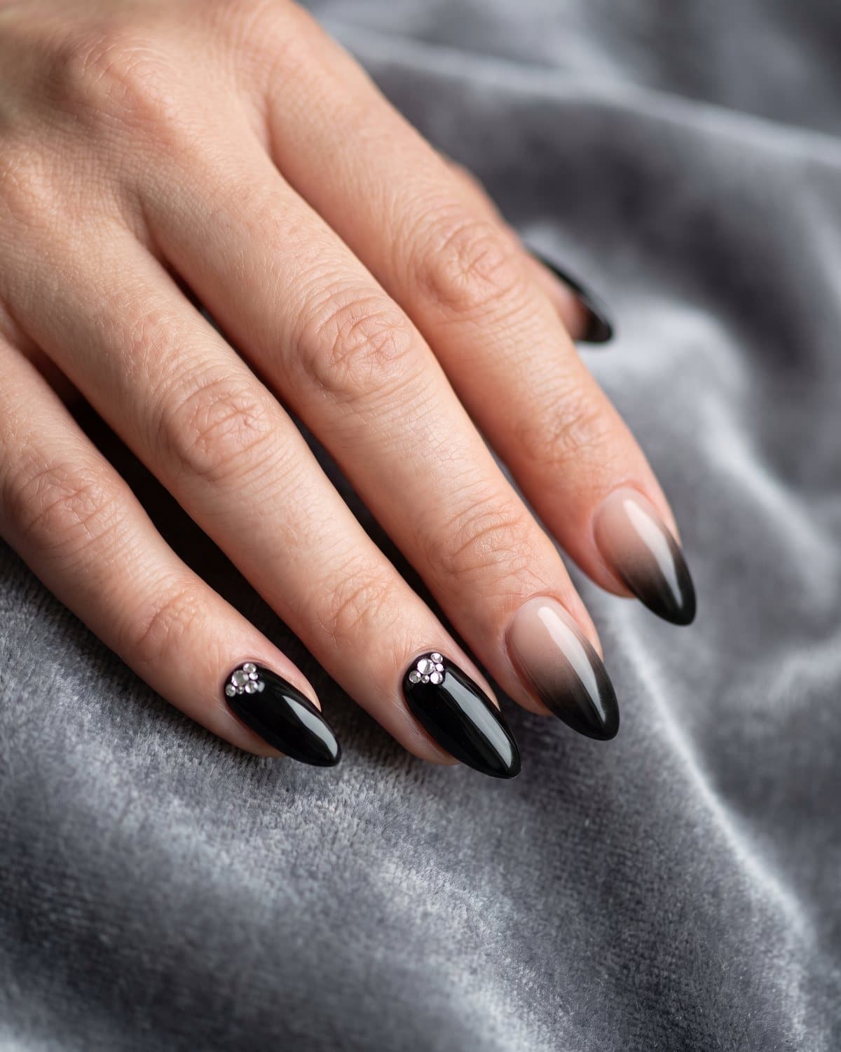 Almond nails in glossy black with a small cluster of clear rhinestones near the cuticle and black-to-nude ombré nails, on grey velvet