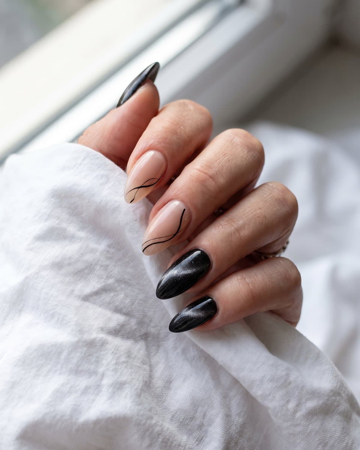 Almond nails with sheer nude and thin black abstract lines and black velvet cat-eye with a soft silver shimmer band, on white cotton