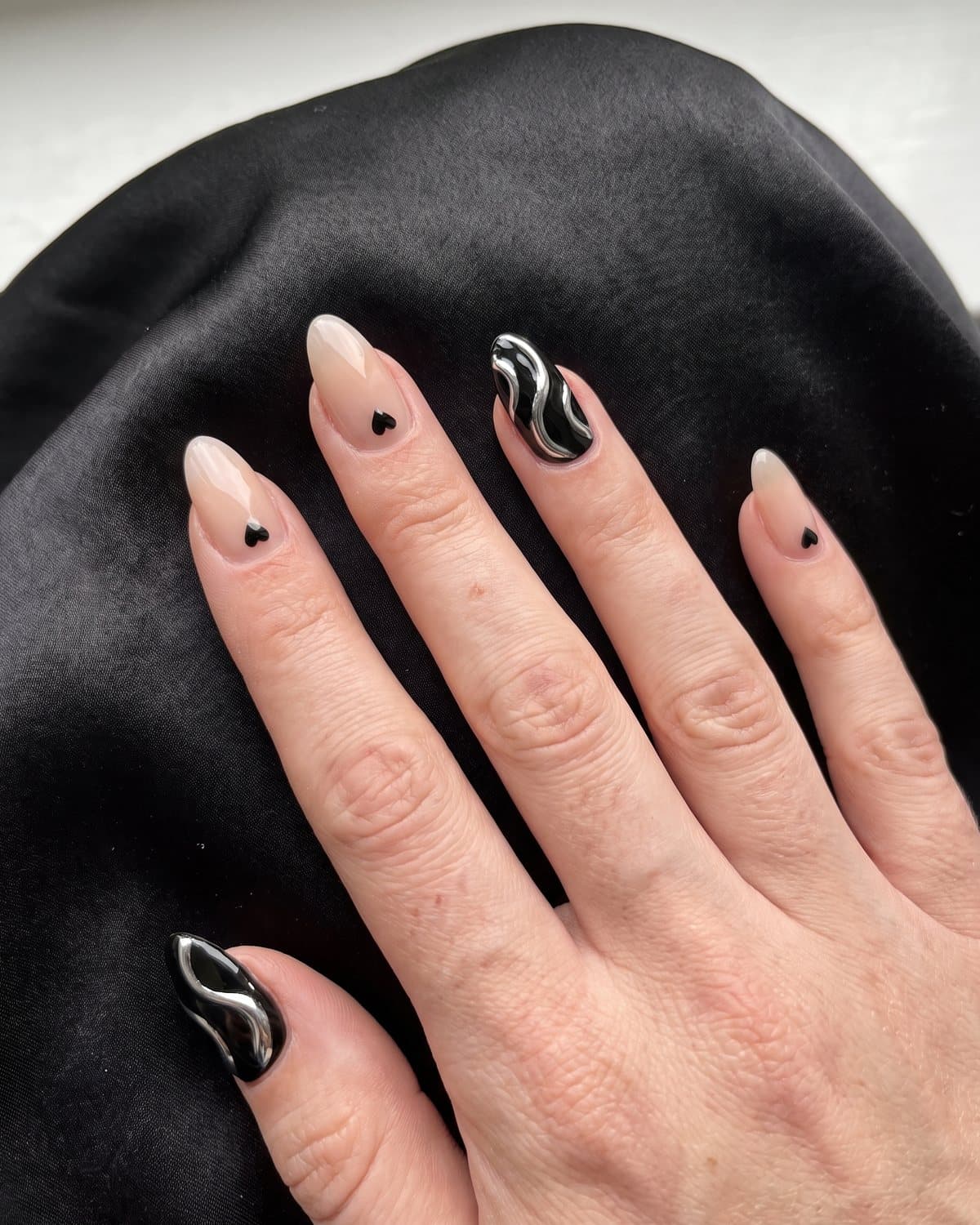 Almond nails in glossy black with flowing silver chrome swirls and sheer nude with a small glossy black heart, on black satin