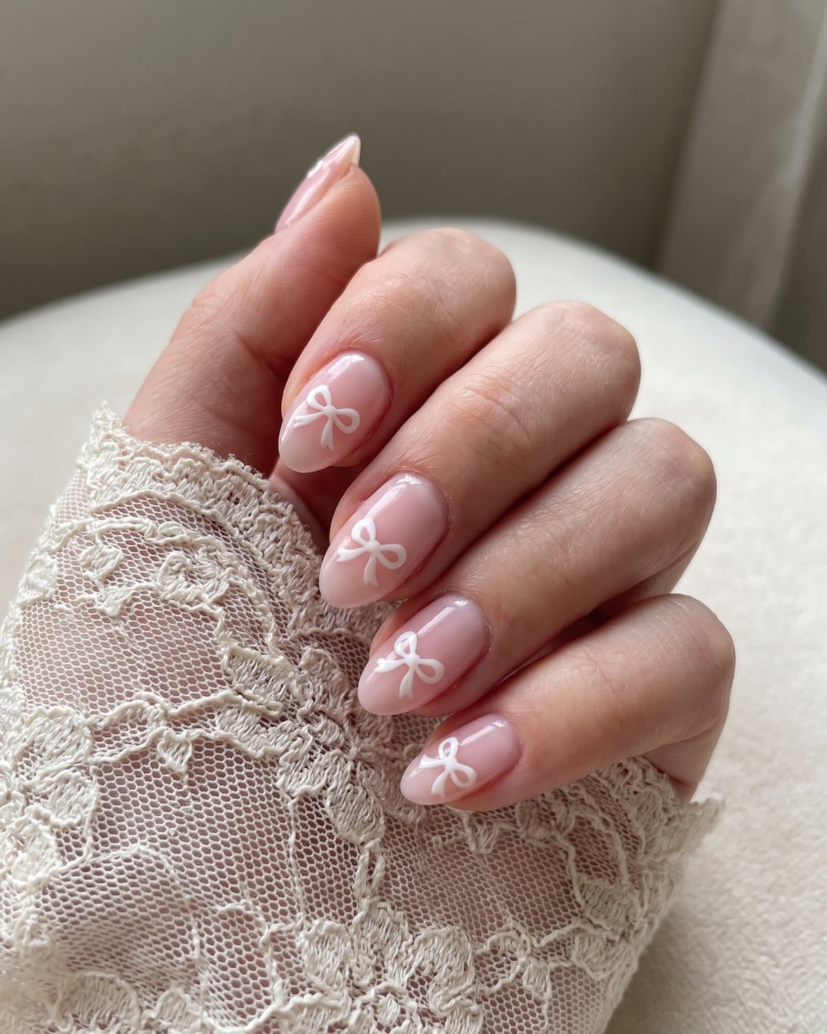Short almond nails in sheer baby pink, each with a tiny hand-painted white bow near the tip, resting on white lace