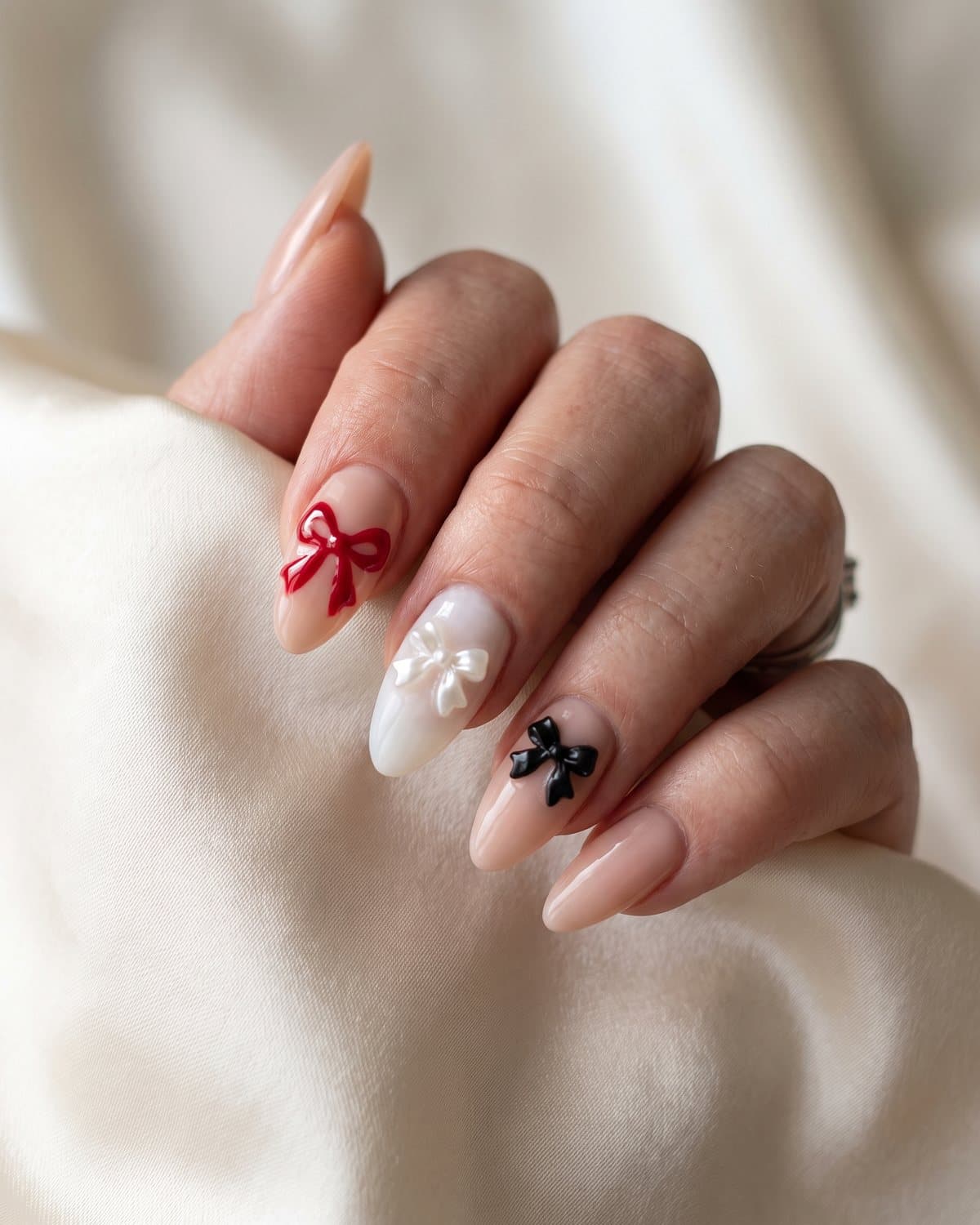 Almond nails with a glossy red painted bow, a milky nail with a 3D pearl bow charm and a nude nail with a small black bow, on cream satin