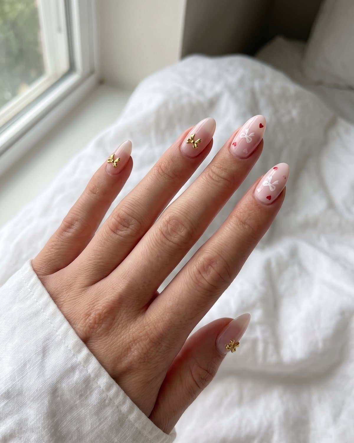 Almond nails with milky pink and a small 3D gold bow charm, and pale pink nails with tiny red hearts and little white bows