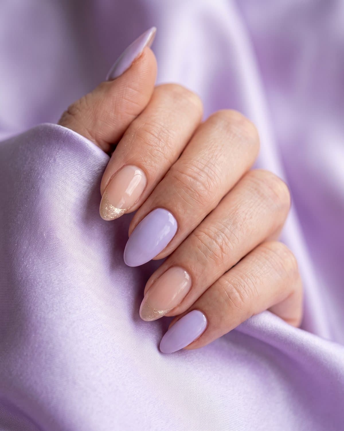 Almond nails in soft lavender and sheer nude with champagne gold shimmer French tips, on lavender satin