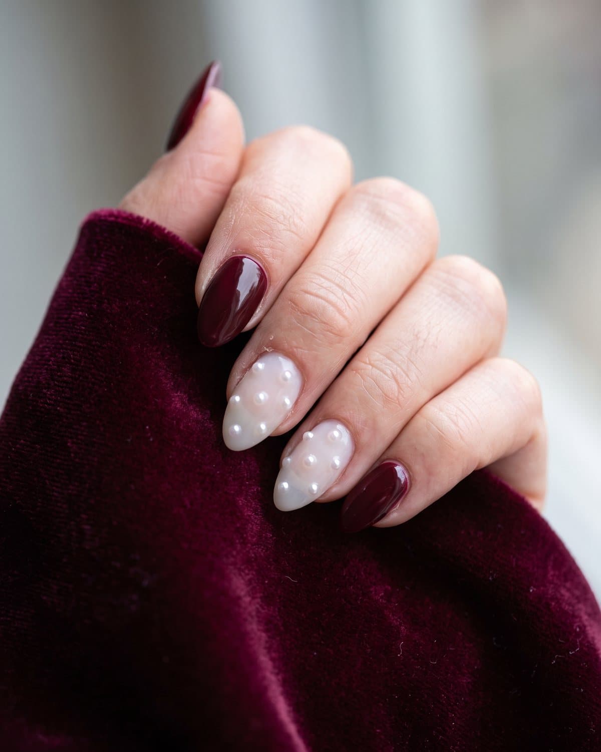 Almond nails in glossy burgundy and milky white with tiny pearl accents, on burgundy velvet