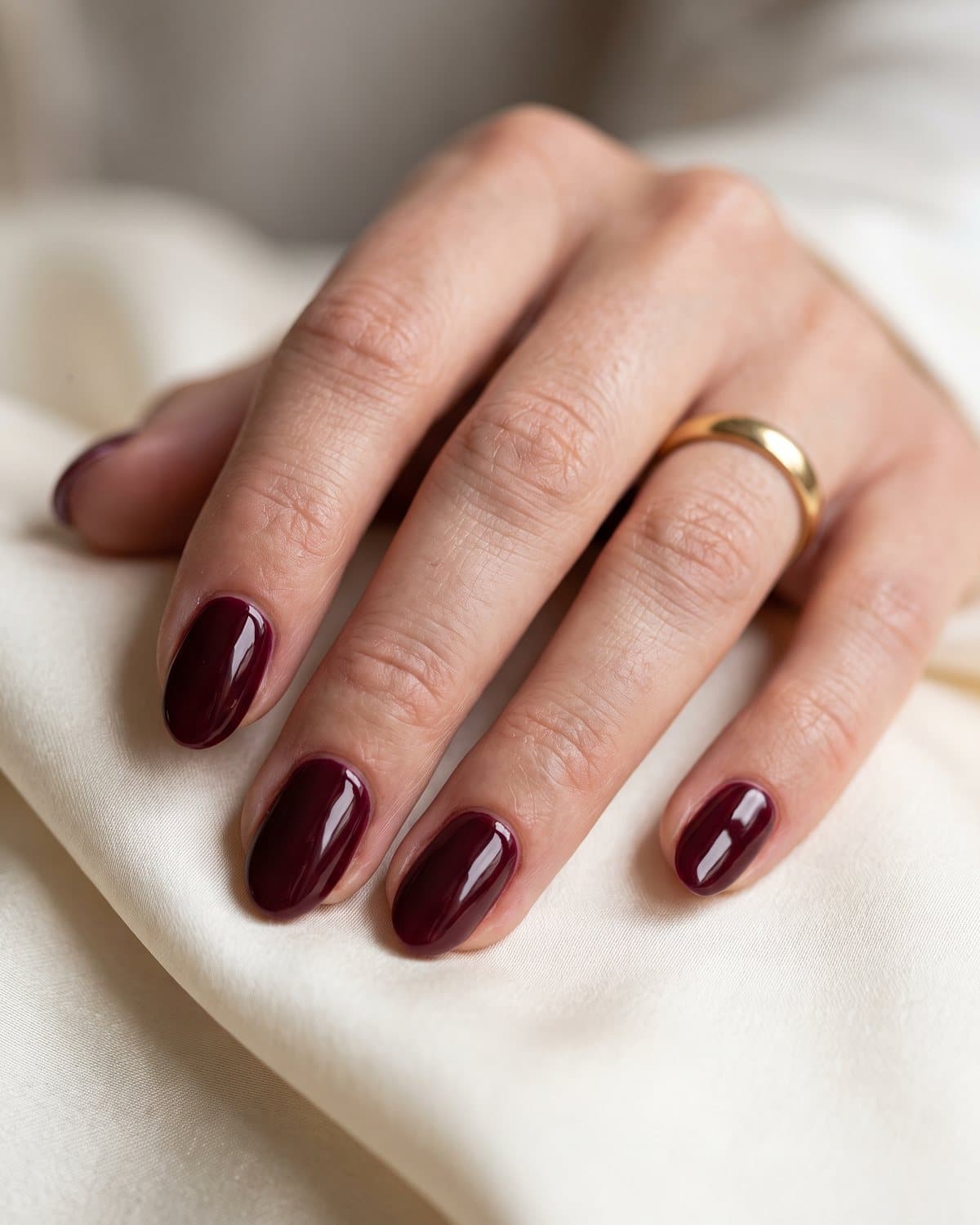 Glossy oval nails in a rich deep burgundy, resting on cream silk with a gold ring