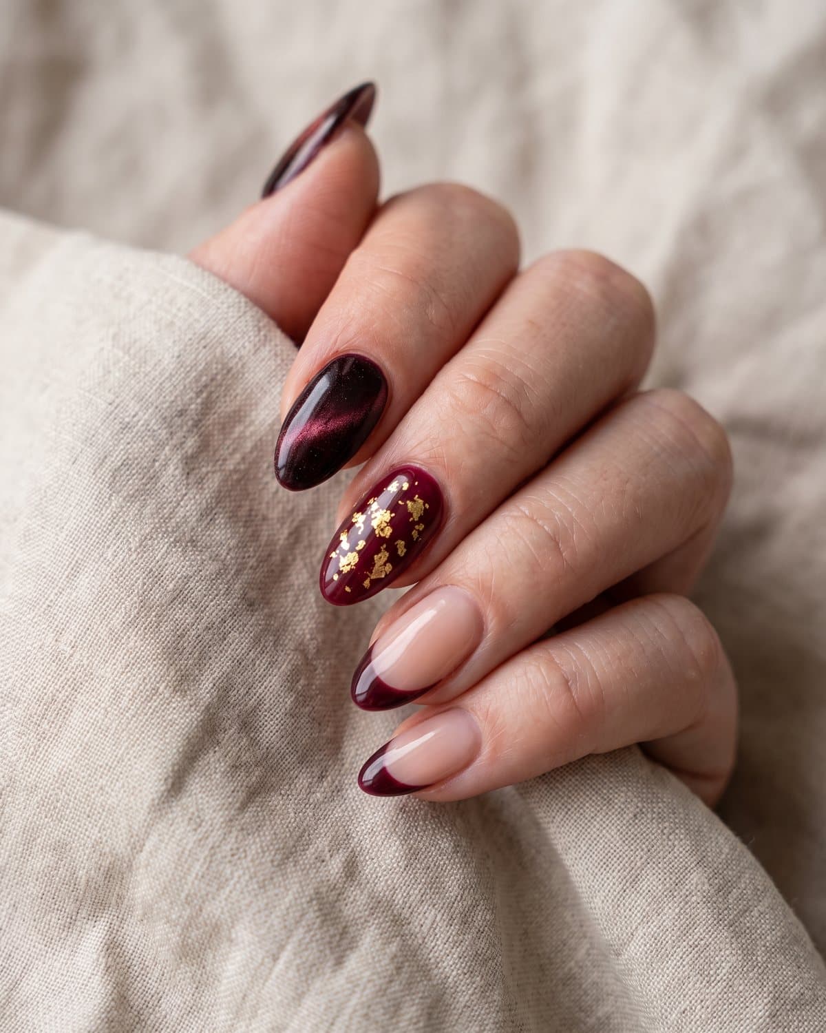 Almond nails with burgundy velvet cat-eye, burgundy with gold foil flakes and sheer nude with burgundy French tips, on beige linen