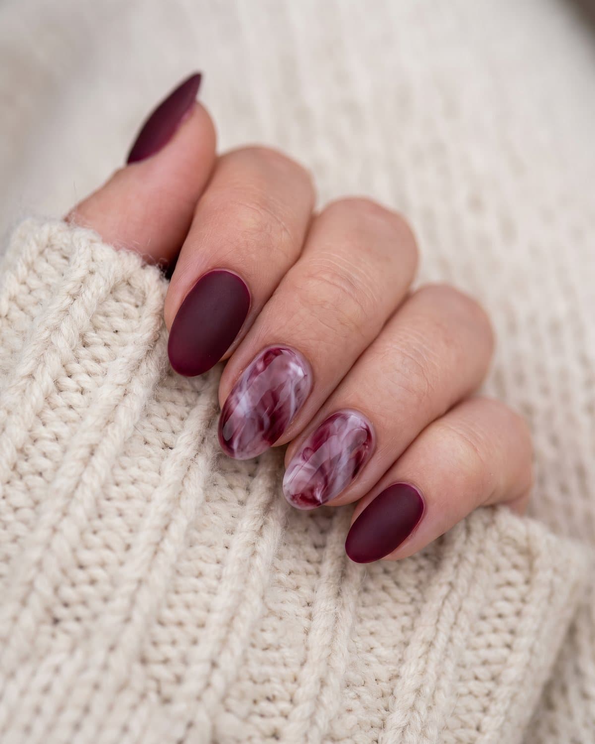 Oval nails with velvety matte burgundy and burgundy and white marble with soft veins, on a cream wool coat