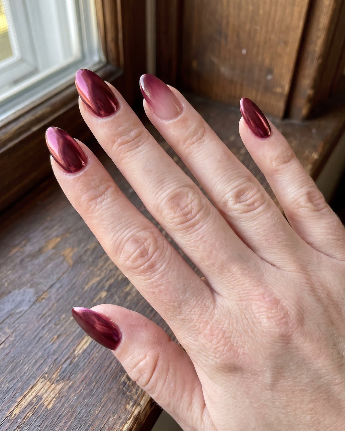 Almond nails with metallic cherry burgundy chrome and nails with a burgundy-to-nude ombré, on dark wood