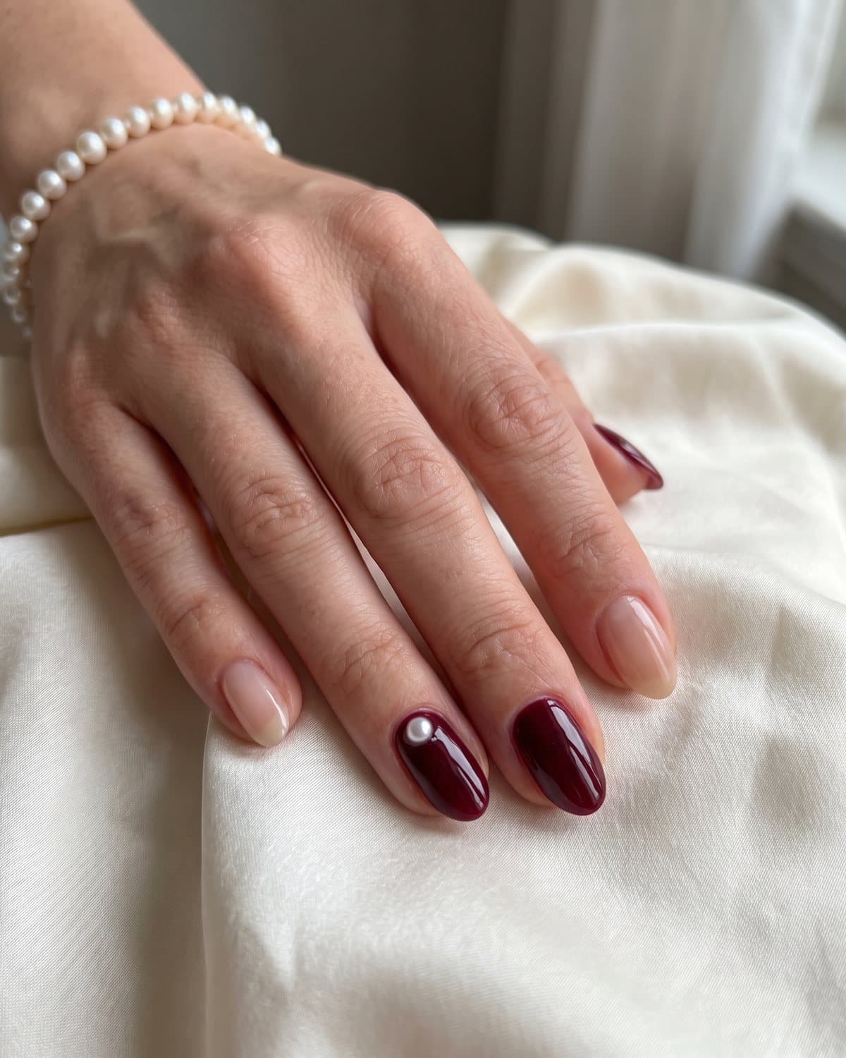 Oval nails in glossy burgundy with a small pearl at the cuticle alternating with sheer nude nails, on ivory silk