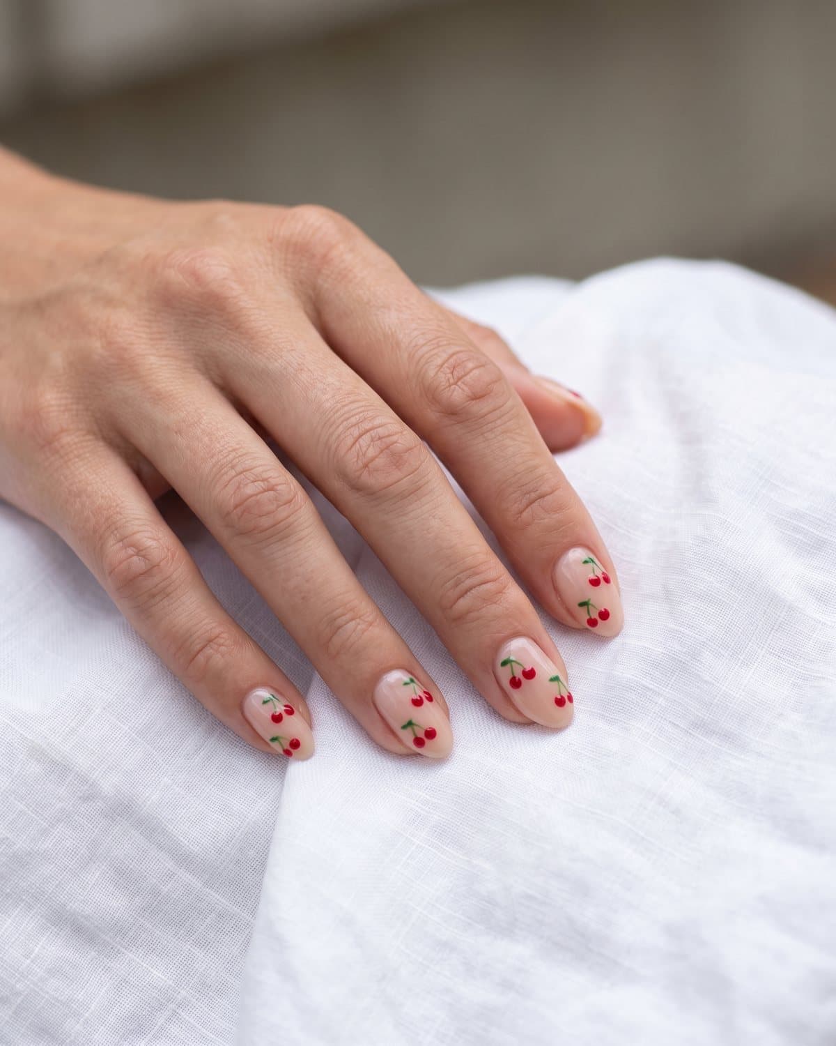 Short almond nails with a sheer nude base and a pair of tiny hand-painted glossy red cherries on each nail, on white linen
