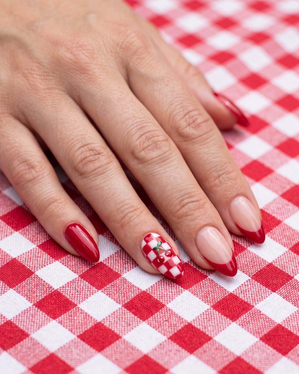 Almond nails with glossy cherry red, sheer nude with cherry red French tips, and a red and white checkerboard nail with a tiny cherry, on red gingham