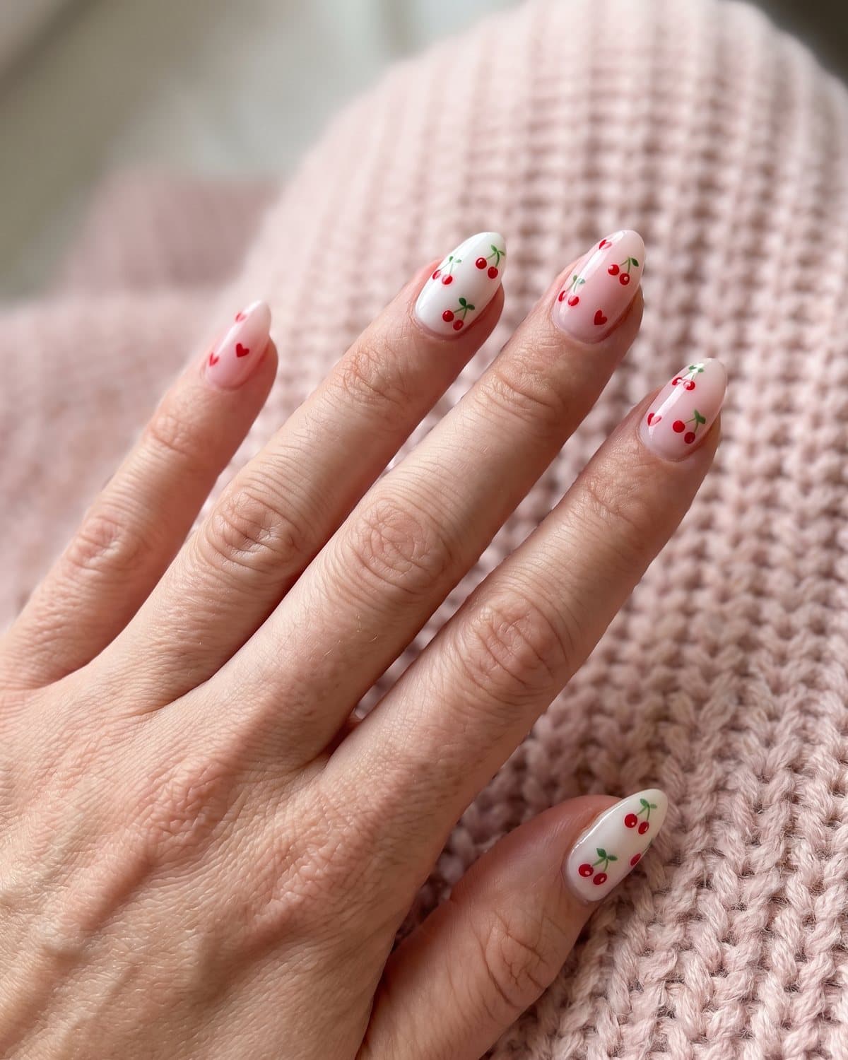 Almond nails with milky white and tiny red cherries, and pale pink nails with small red hearts and cherries, on a pink knit