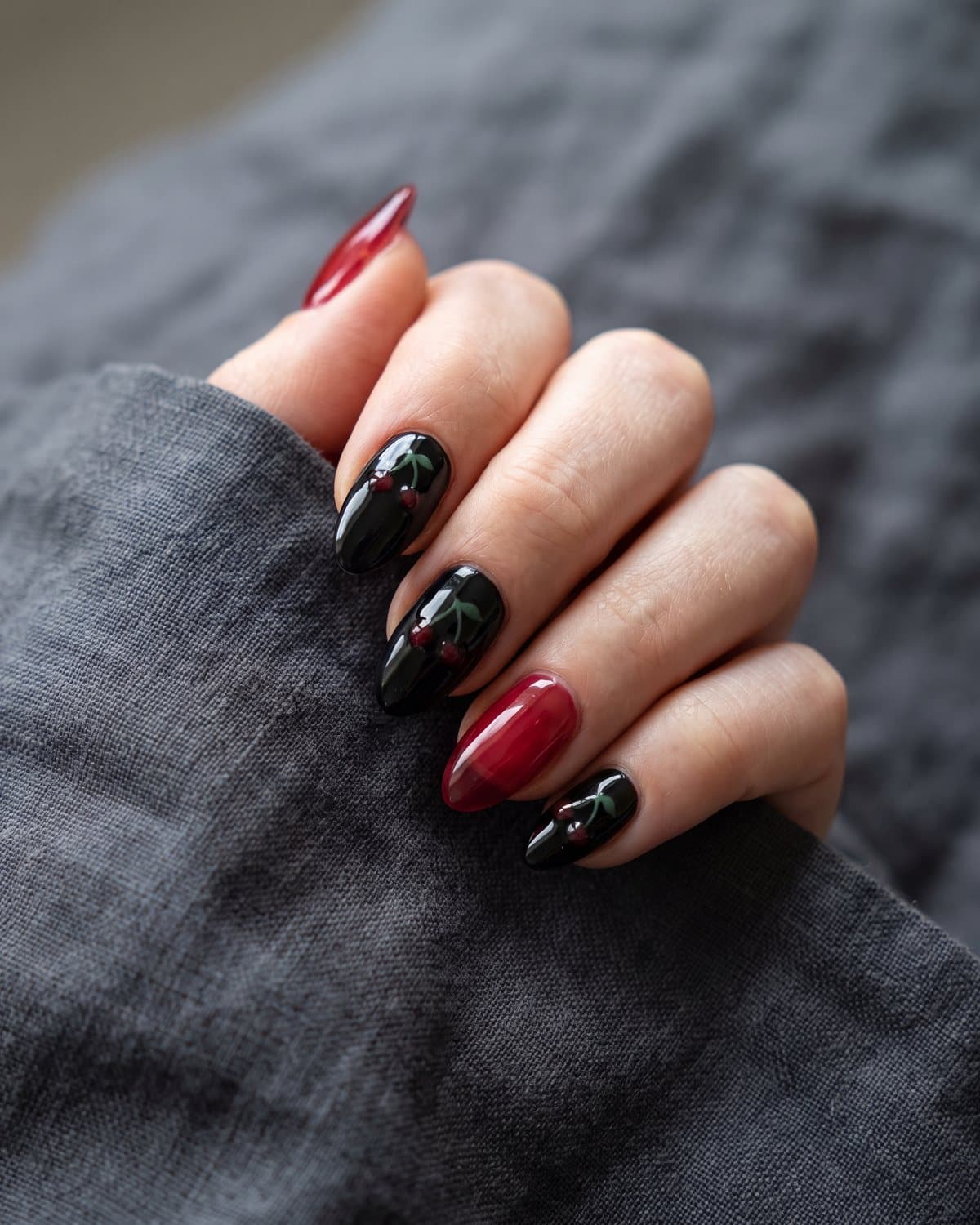 Almond nails in glossy black with small dark red cherries and translucent cherry red jelly nails, on dark grey linen