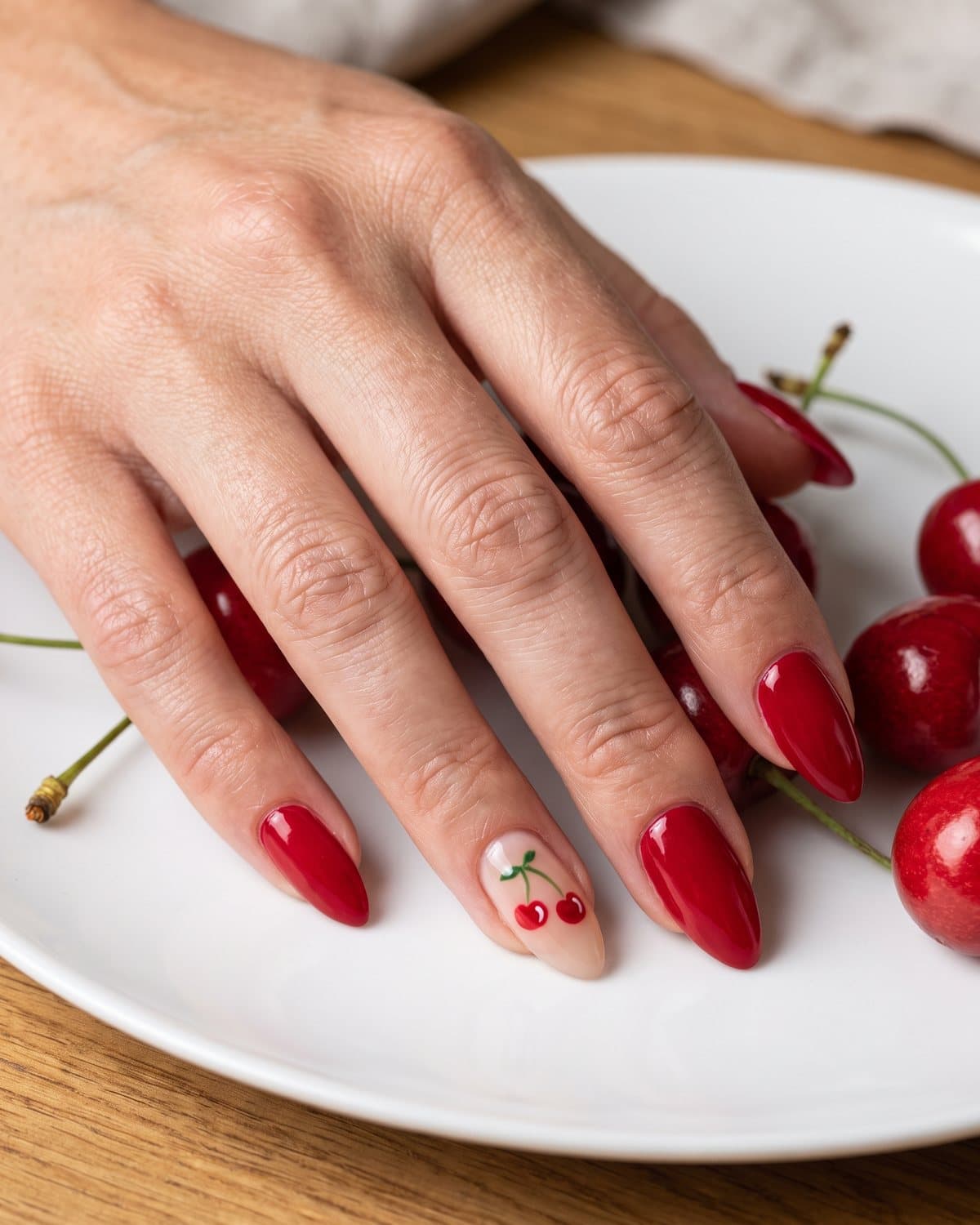 Almond nails with tiny hand-painted red cherries on a sheer nude base and glossy cherry red nails, beside fresh cherries on a white plate