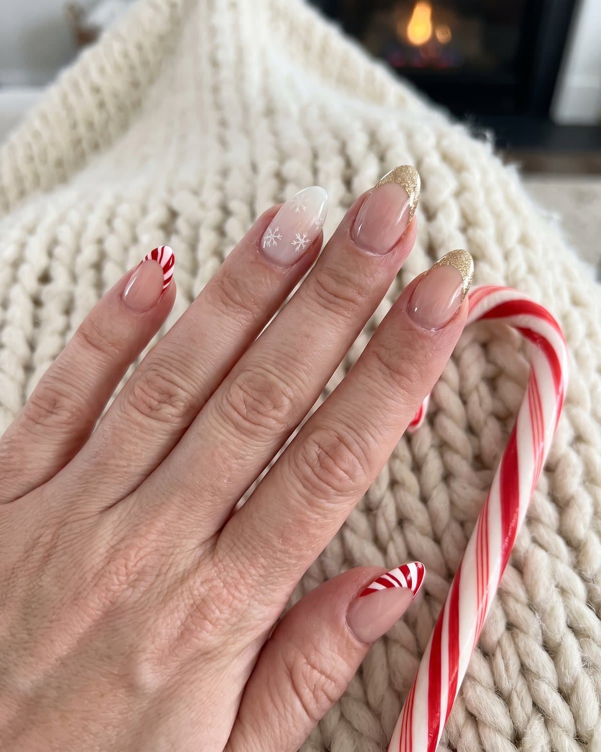 Almond nails with red and white candy cane French tips, gold glitter French tips and white snowflakes on milky white, on a cream knit blanket