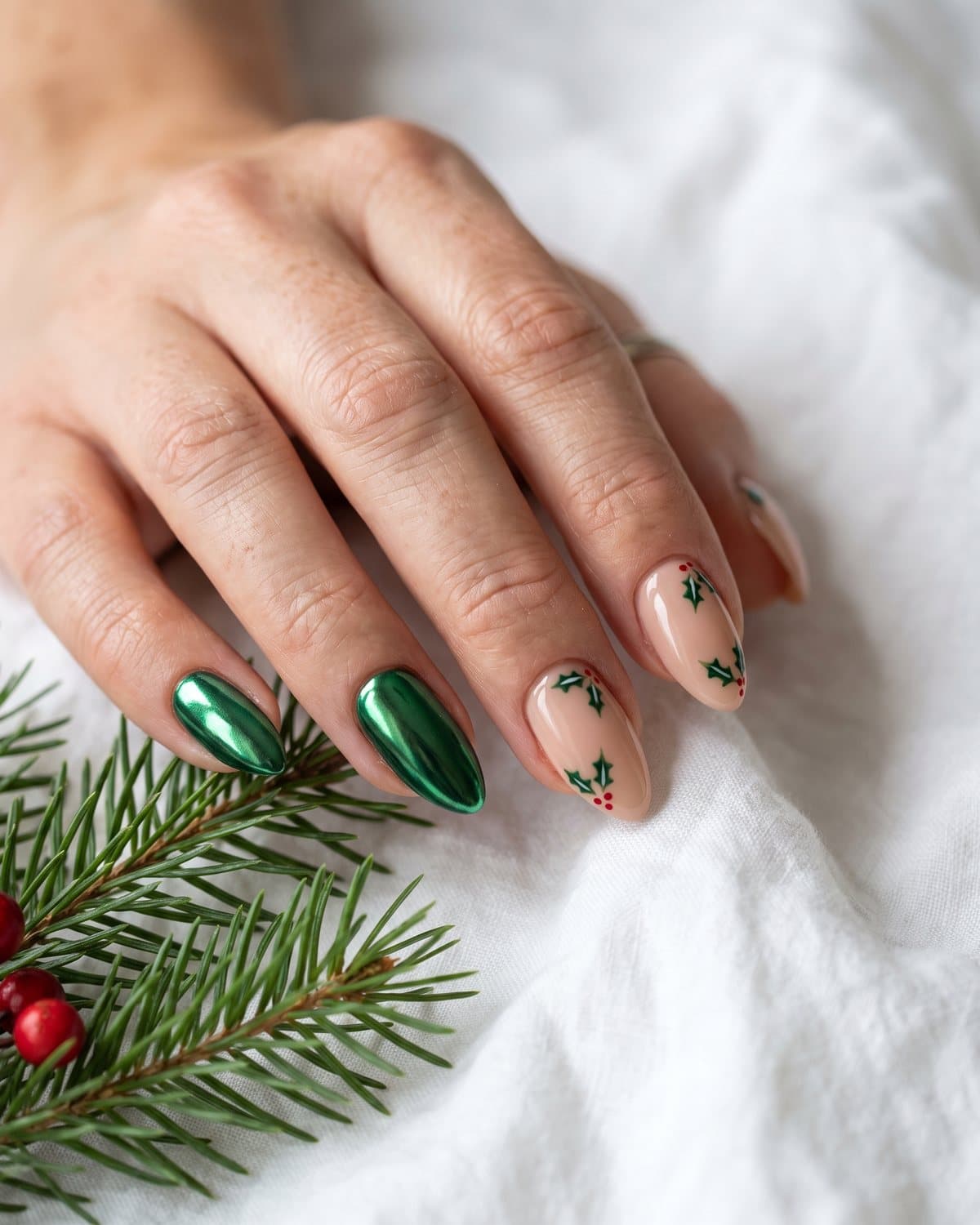 Almond nails in metallic emerald chrome and nude nails with hand-painted holly leaves and red berries, beside pine branches