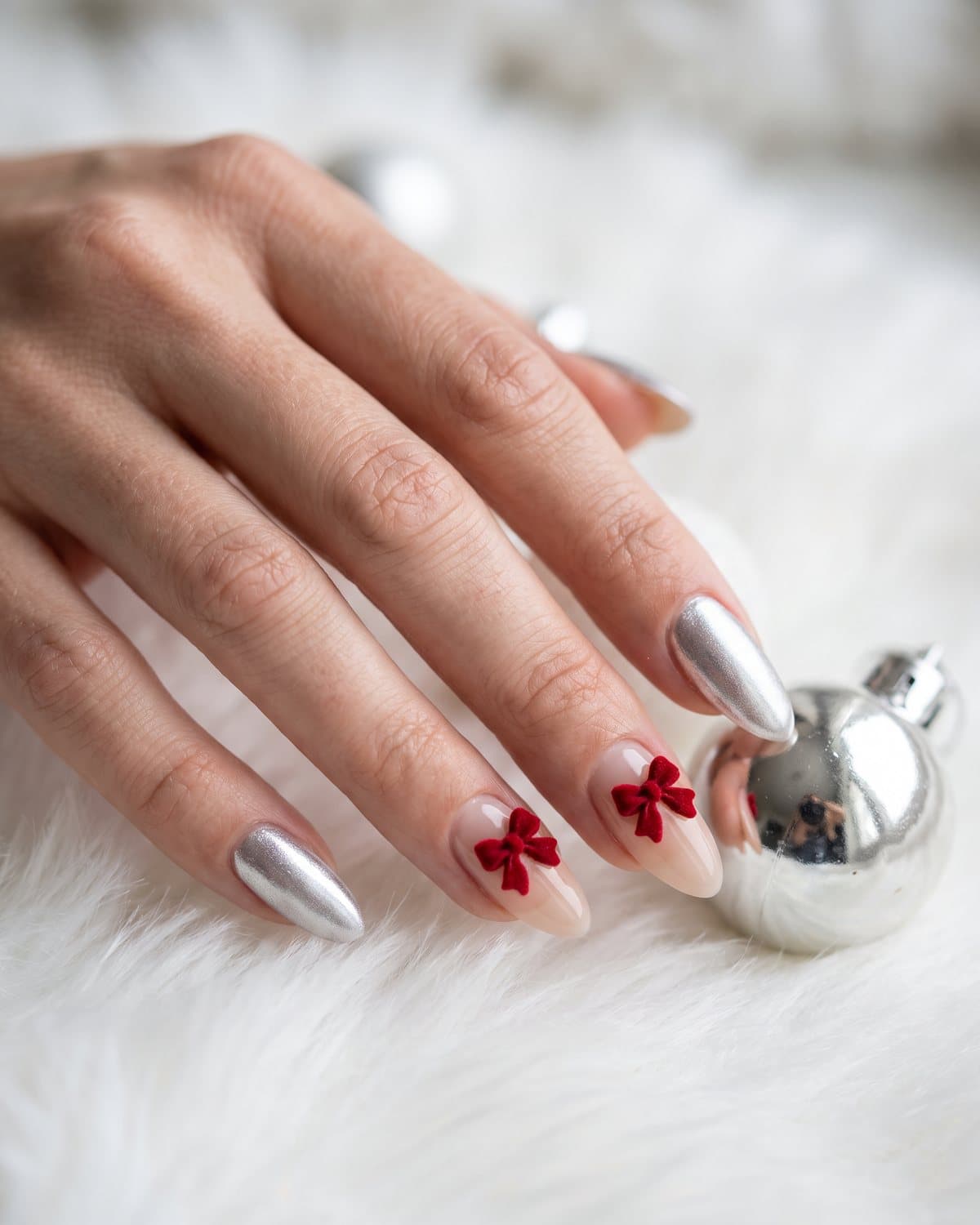 Almond nails with tiny red bows on a sheer nude base and frosted silver chrome nails, on white faux fur next to a silver ornament