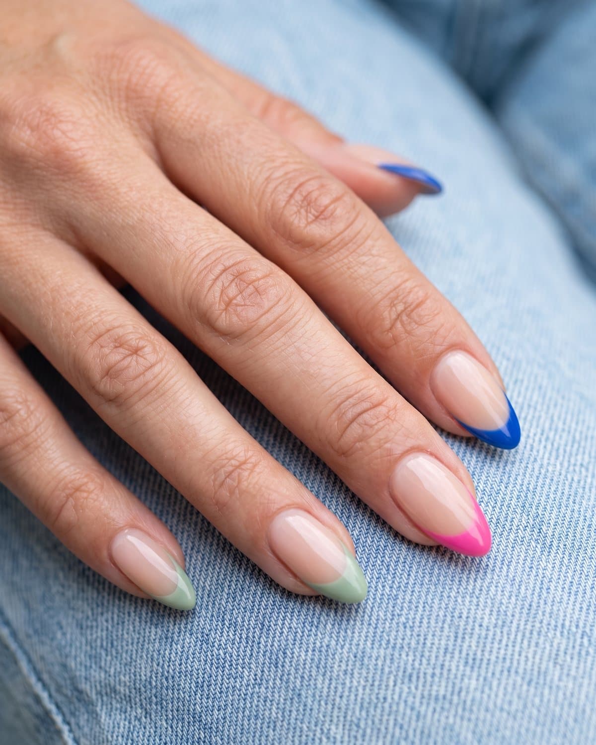 Almond nails with a sheer nude base and French tips in cobalt blue, hot pink and sage green, resting on denim