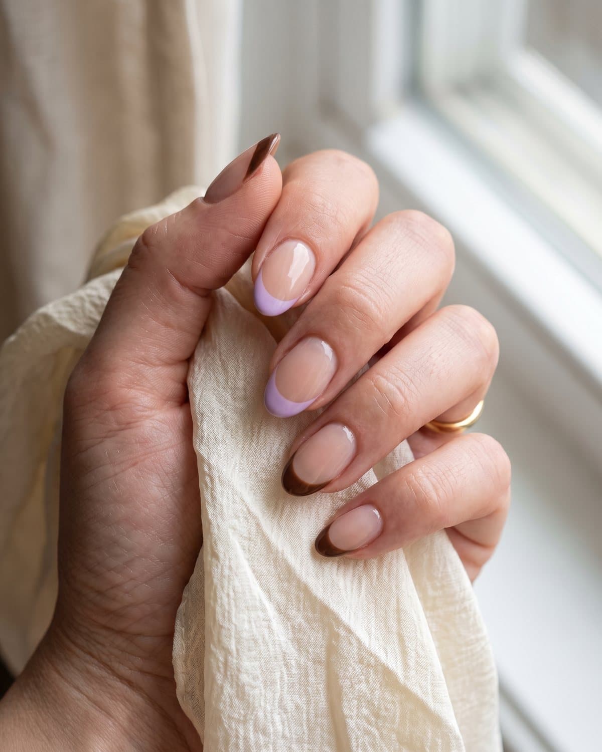Oval nails with a sheer nude base and French tips alternating between soft lilac and chocolate brown, on a cream silk scarf