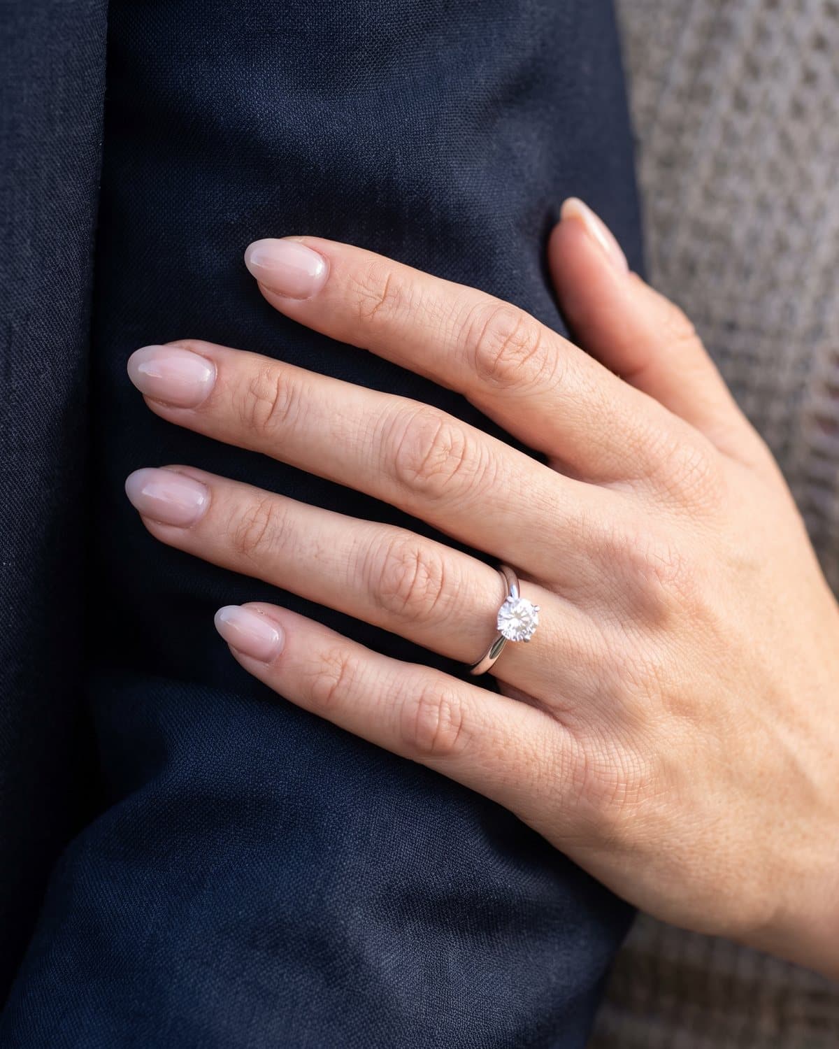 Short almond nails in glossy sheer pink wearing a solitaire diamond engagement ring, resting on a dark suit sleeve