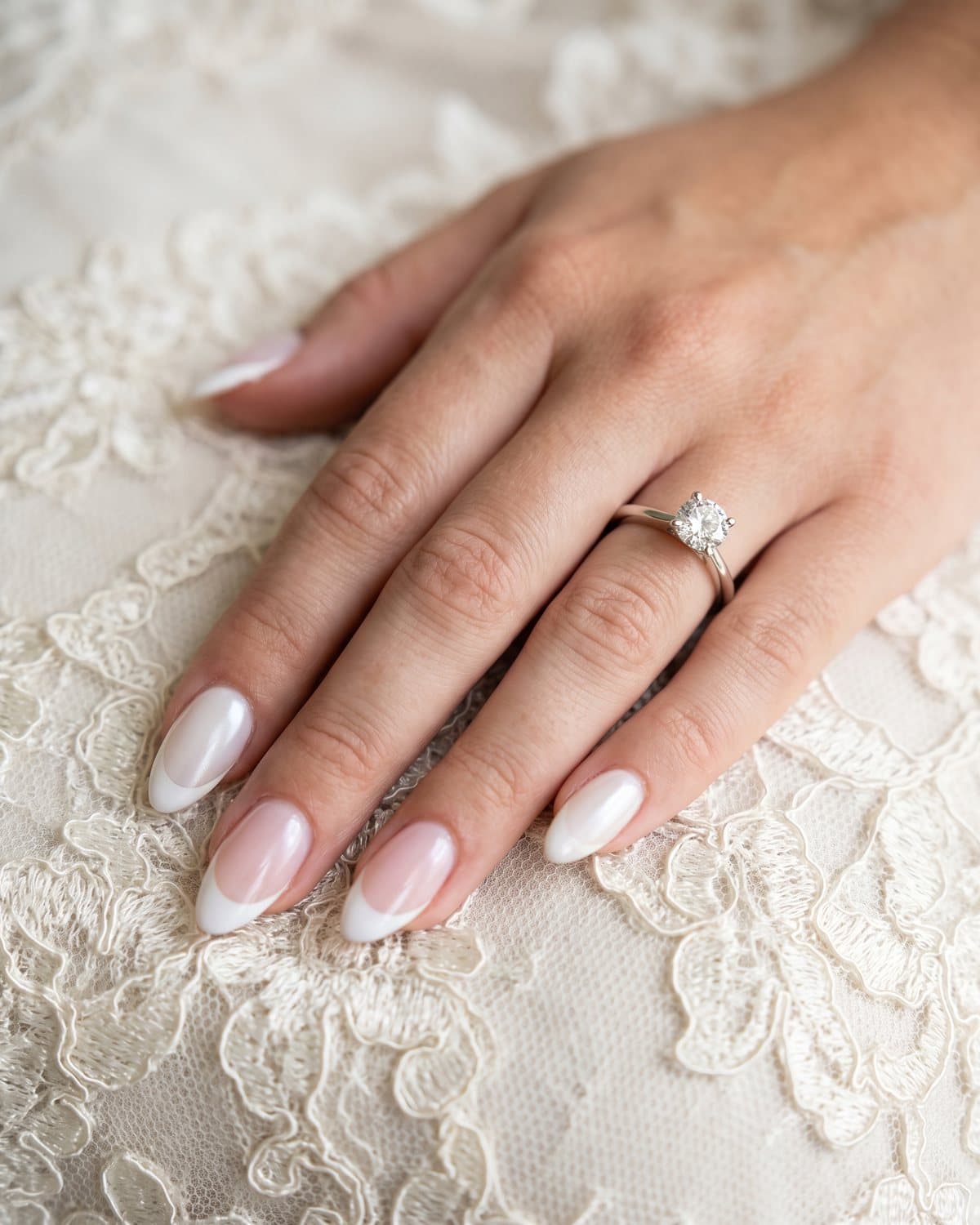 Almond nails with milky white, classic white French tips on sheer pink and a soft pearl glaze, with a diamond engagement ring on ivory lace