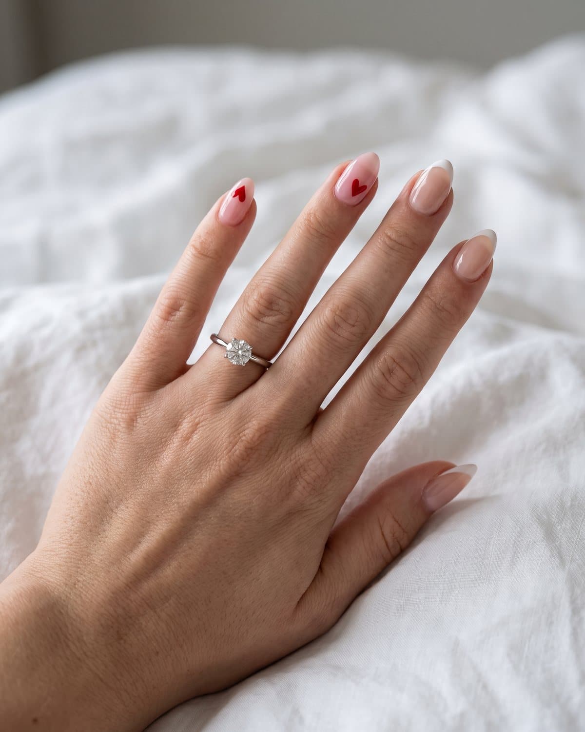 Almond nails with sheer pink and a tiny red heart accent and sheer nude with ultra-thin micro white French tips, with a diamond engagement ring