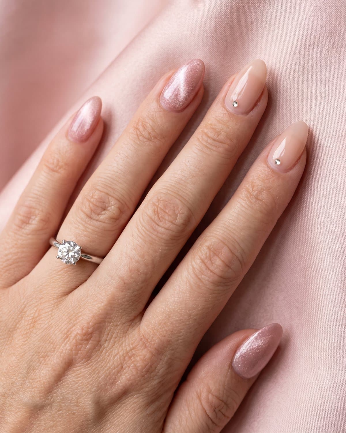 Almond nails with blush pink cat-eye shimmer and sheer nude with a single small rhinestone near the cuticle, with a diamond engagement ring on blush satin