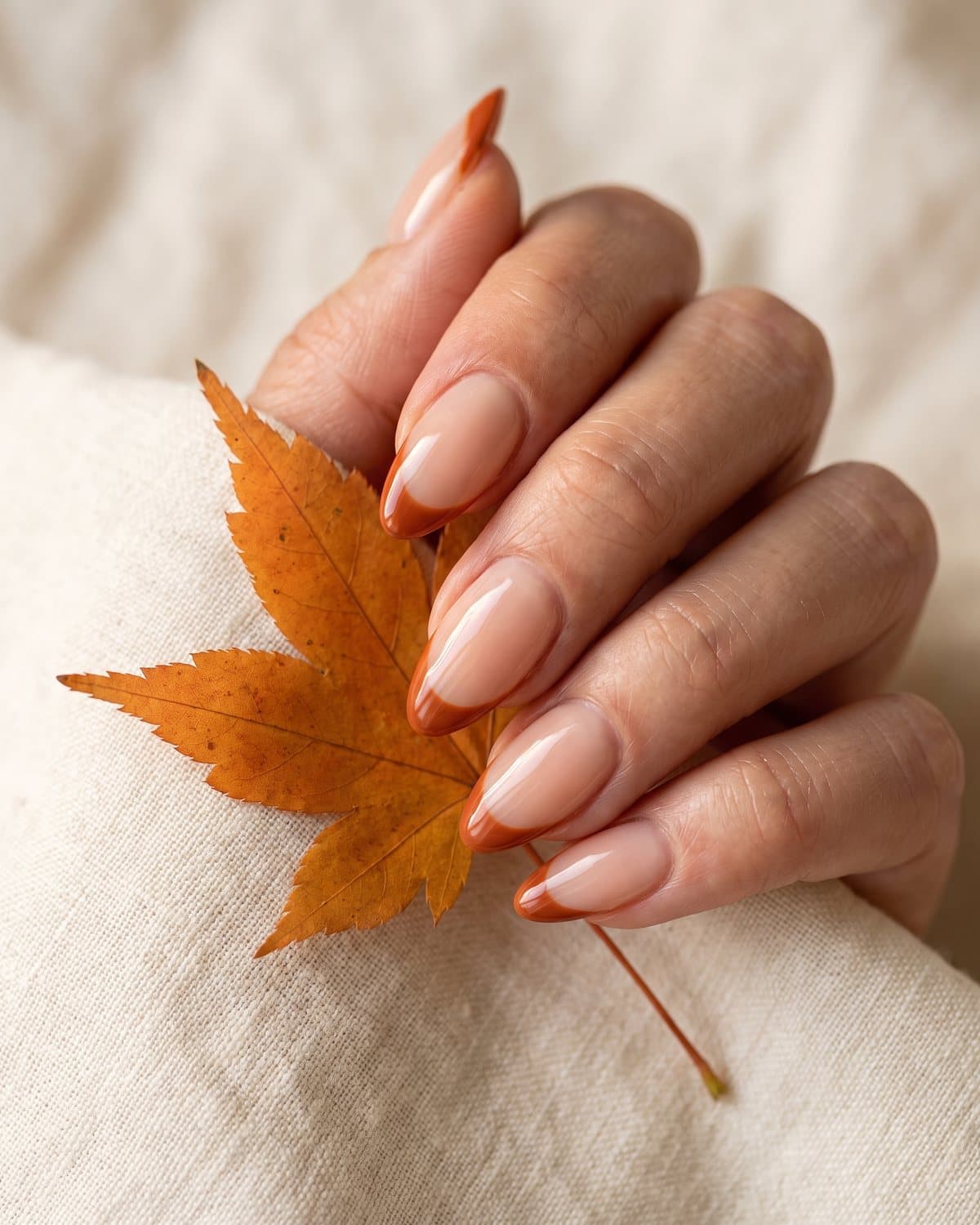 Almond nails with a sheer nude base and burnt orange French tips, holding a single orange maple leaf