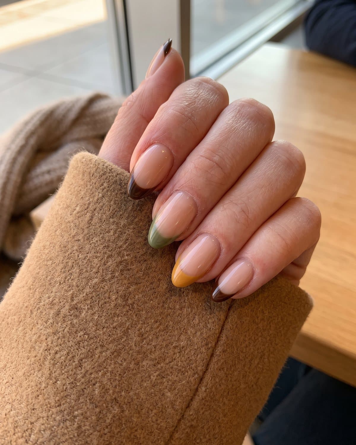 Almond nails with a sheer nude base and French tips alternating chocolate brown, olive green and mustard yellow, on a camel wool coat
