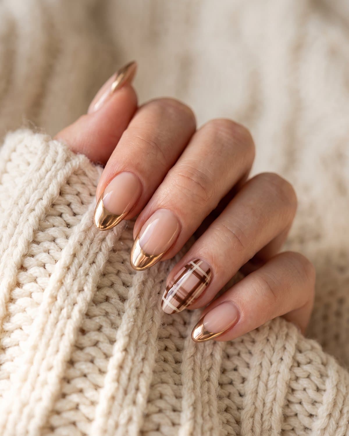 Almond nails with mirror gold chrome French tips and brown and cream plaid French tips, on a cream knit blanket