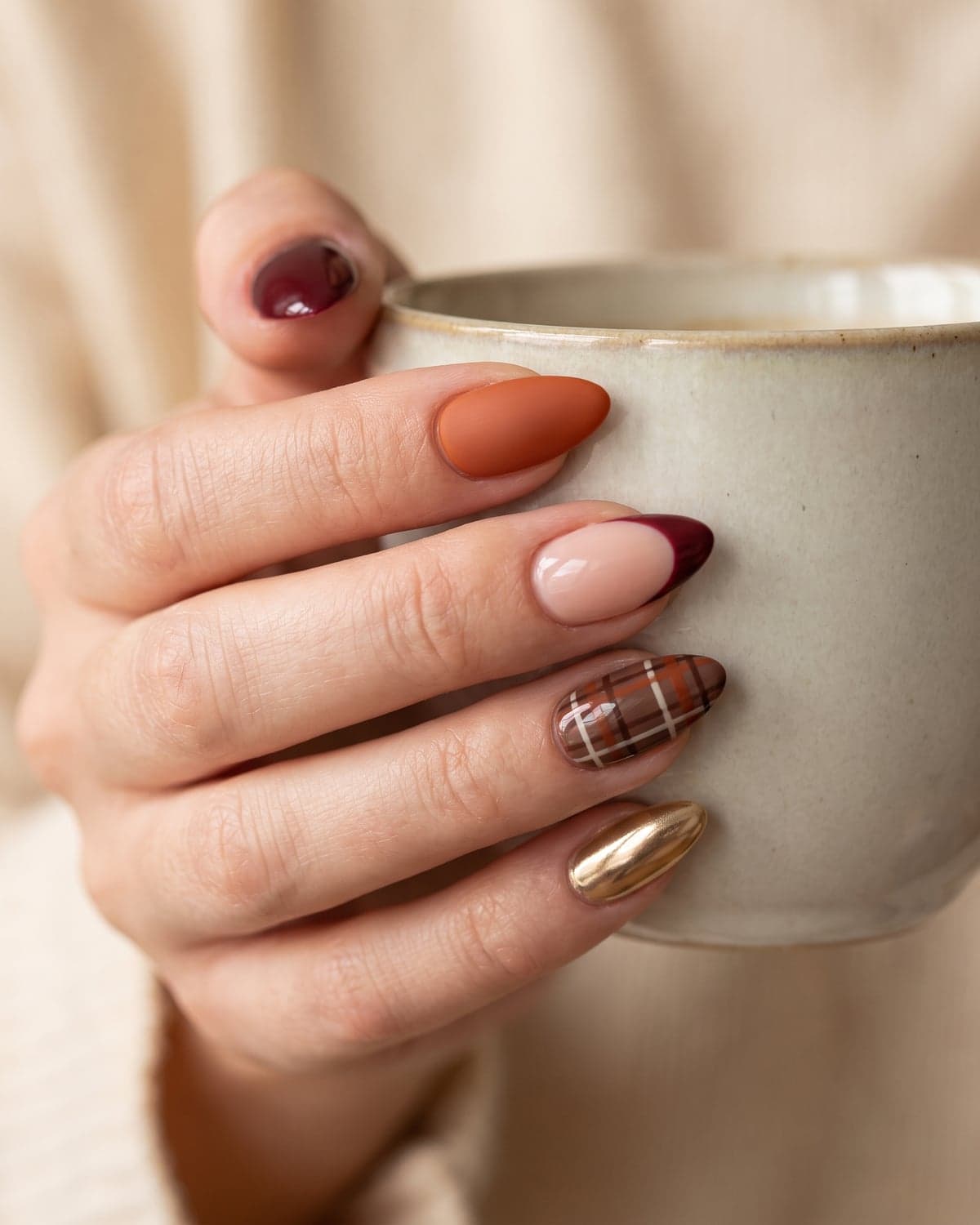 Burgundy, burnt orange and nude almond nails with a plaid accent and gold chrome nail on a warm cream background