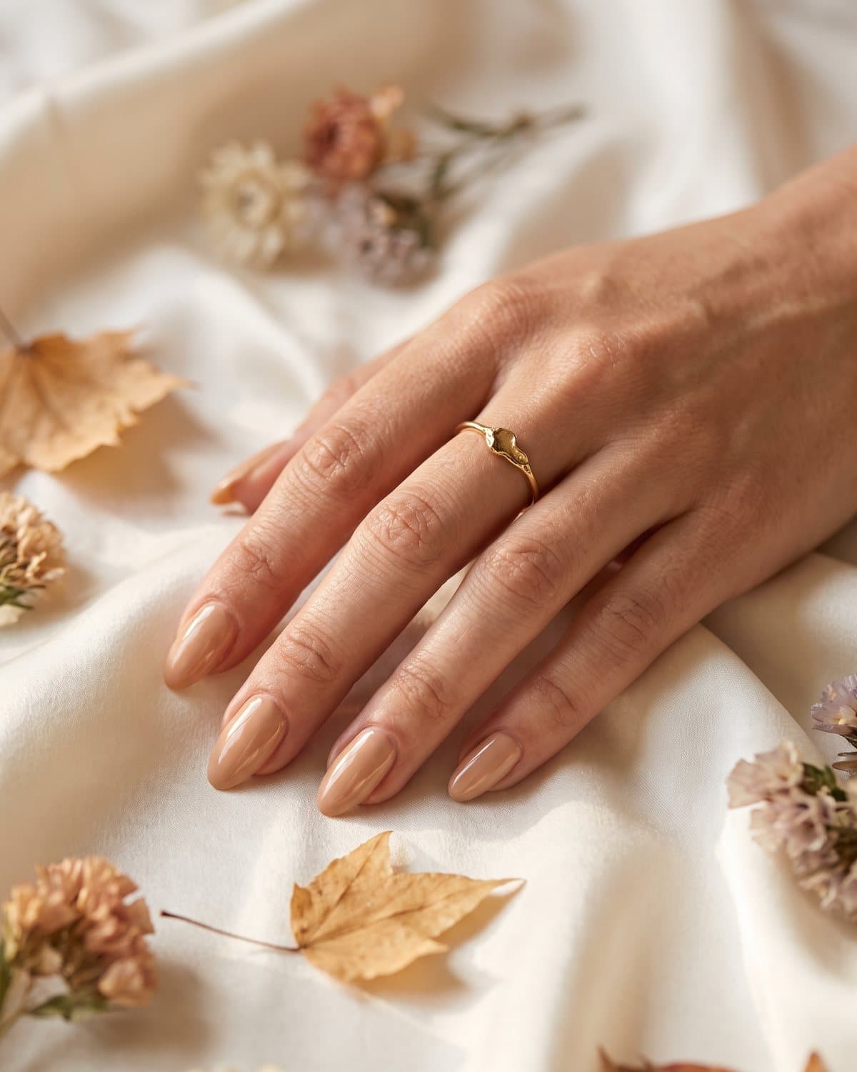 Glossy almond nails in a warm champagne nude with a delicate gold ring, resting on ivory silk with dried autumn flowers