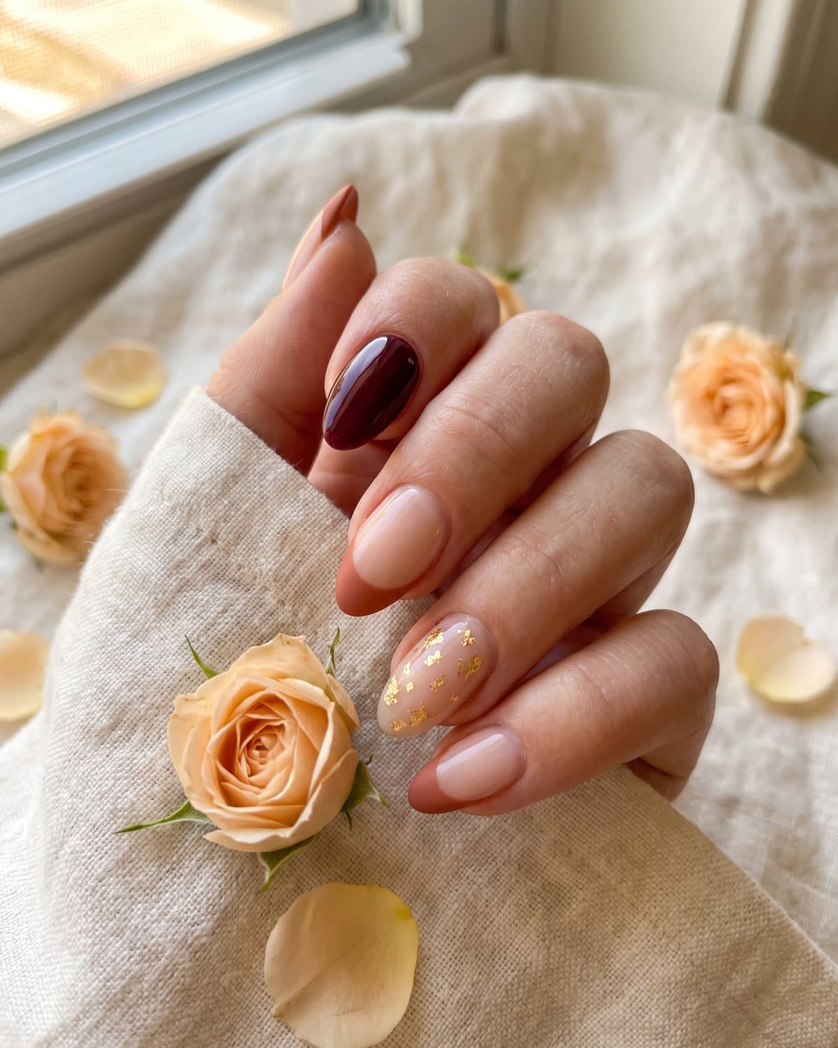 Almond nails with glossy burgundy, sheer nude with terracotta French tips and a nude nail with gold leaf flakes, on cream linen with small orange roses