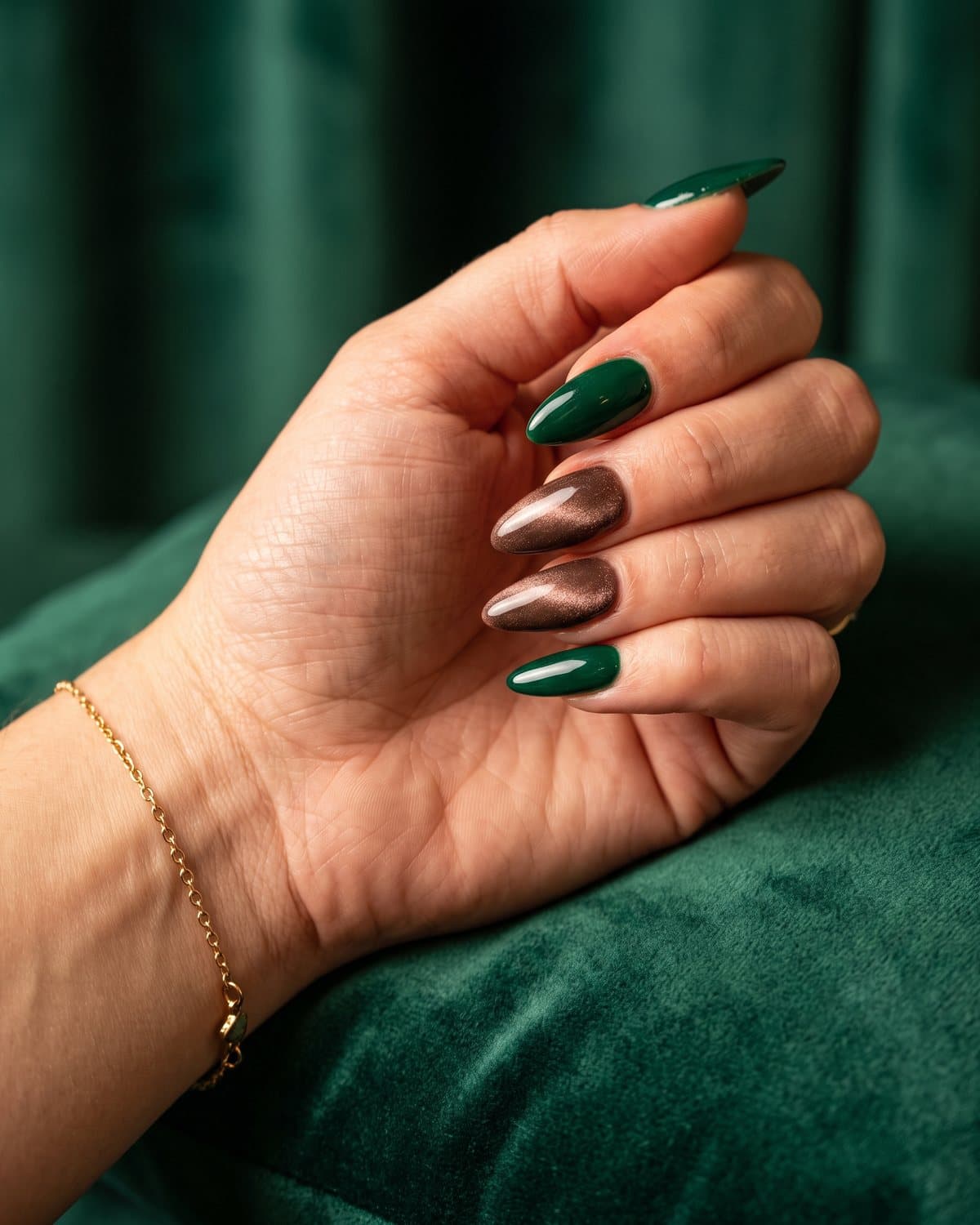 Almond nails in glossy deep emerald green and mocha brown velvet cat-eye, on emerald velvet with a gold bracelet