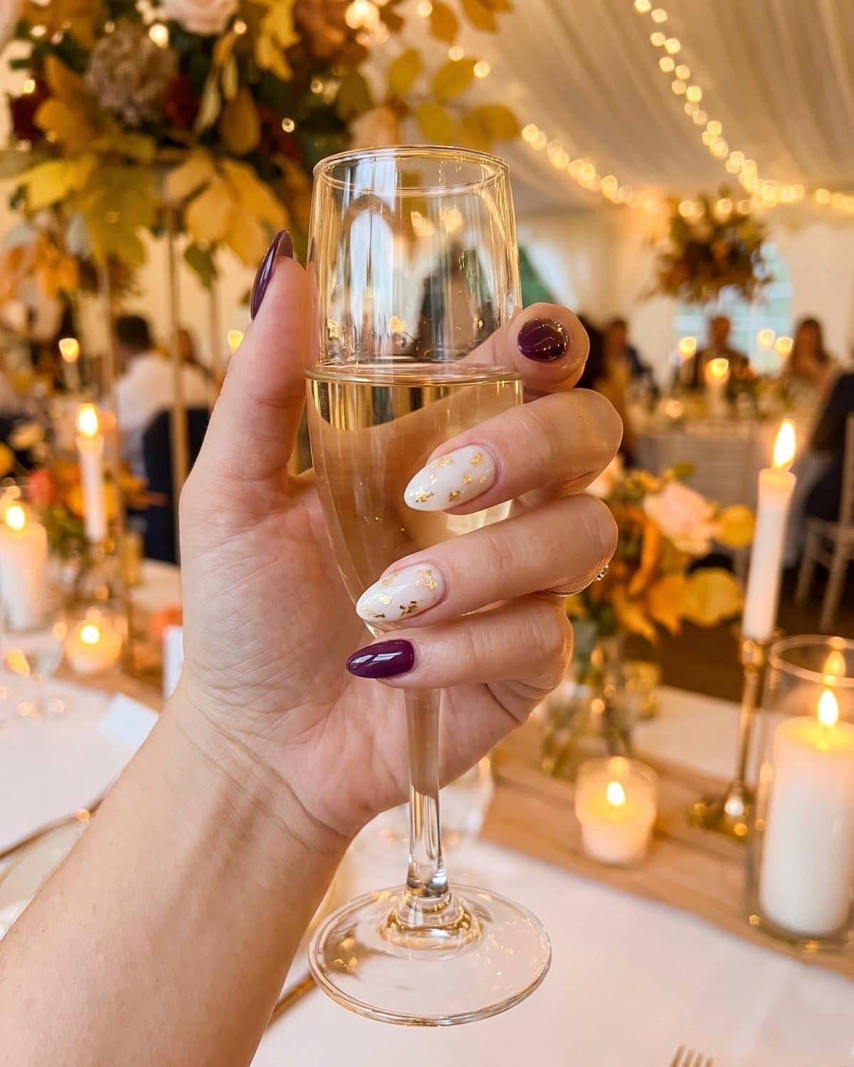Almond nails in glossy deep plum and milky white with fine gold leaf, holding a champagne glass at a candlelit autumn wedding reception