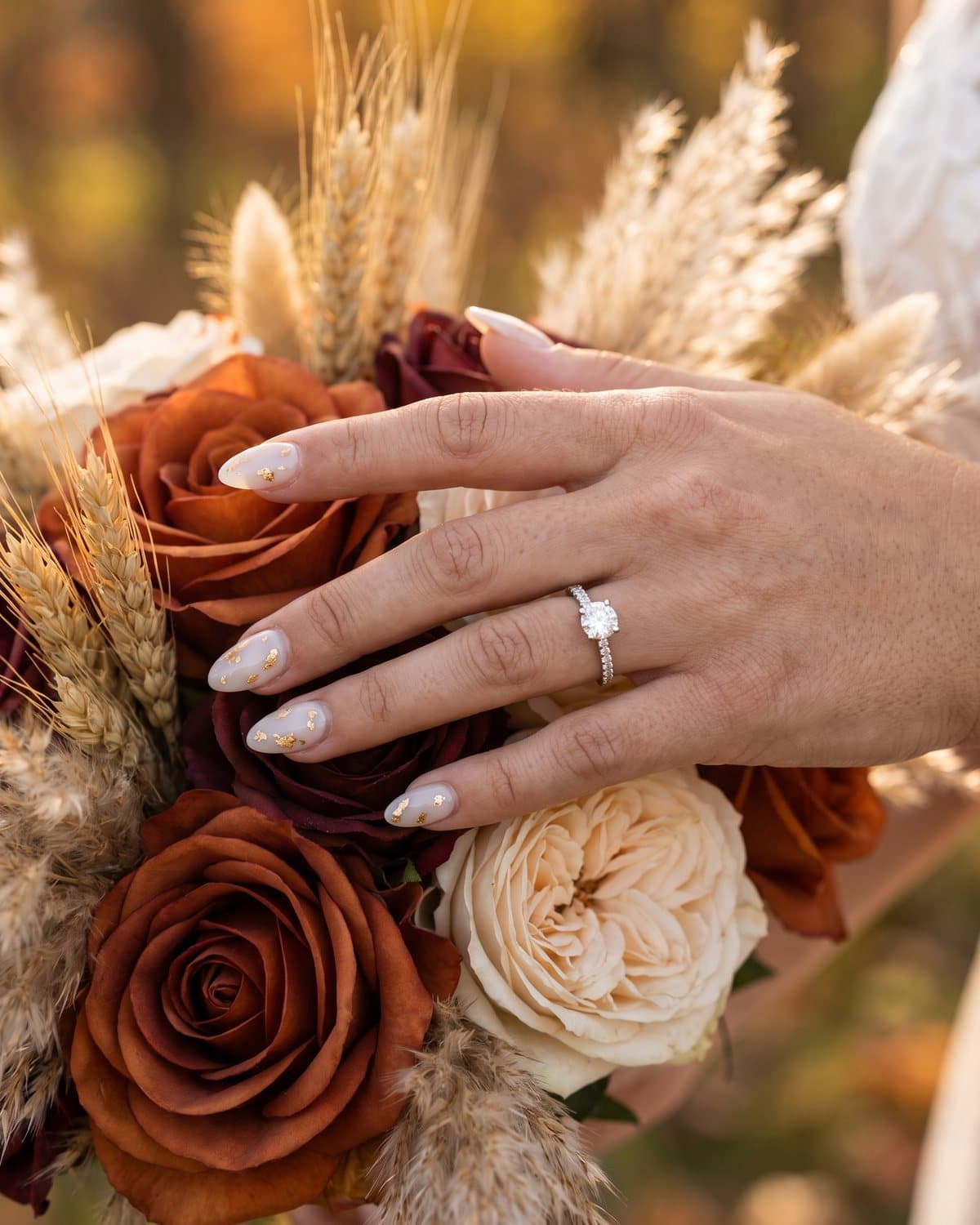 Bride's hand with an engagement ring and milky white almond nails with gold leaf, holding a bouquet of burnt orange, burgundy and cream roses