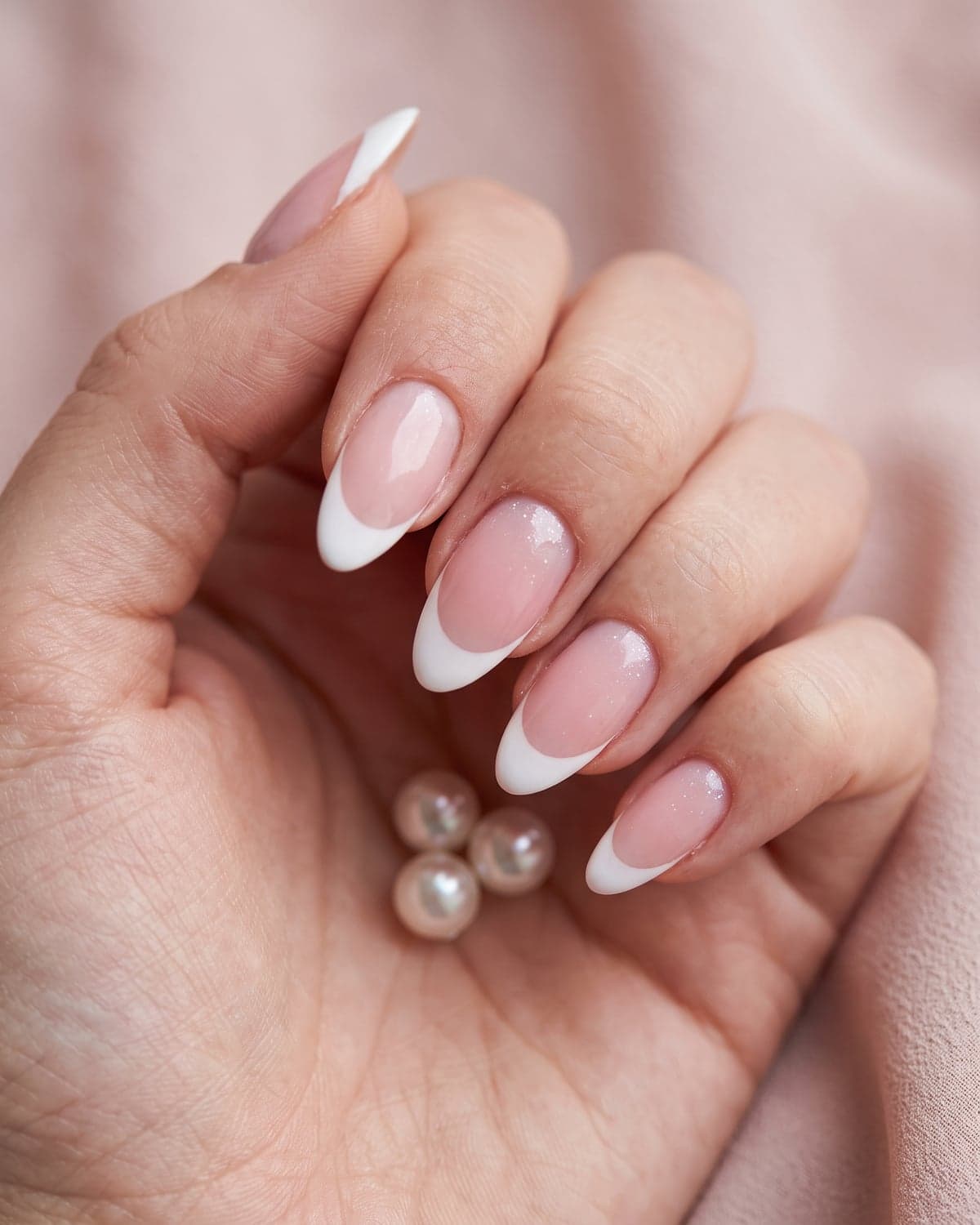 Classic white French tip almond nails on a sheer pink base with a soft blush background and pearls
