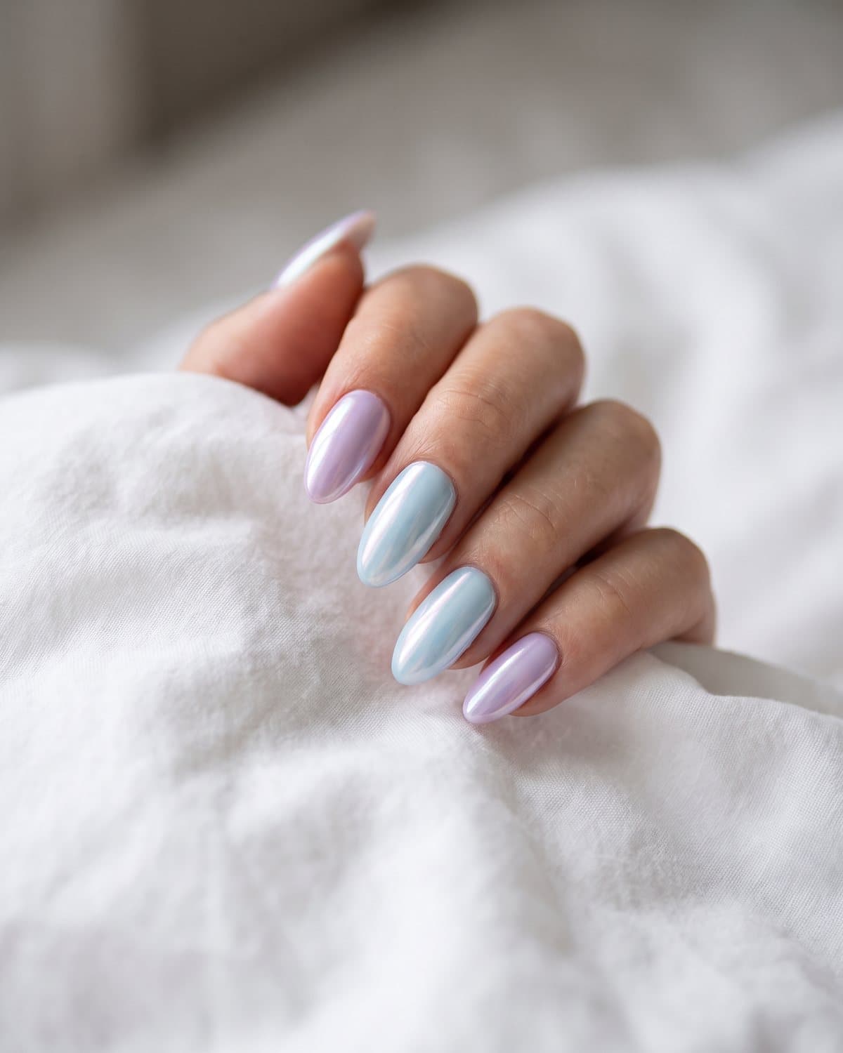 Almond nails alternating between pale lavender glazed and baby blue blueberry glazed, resting on white bedding