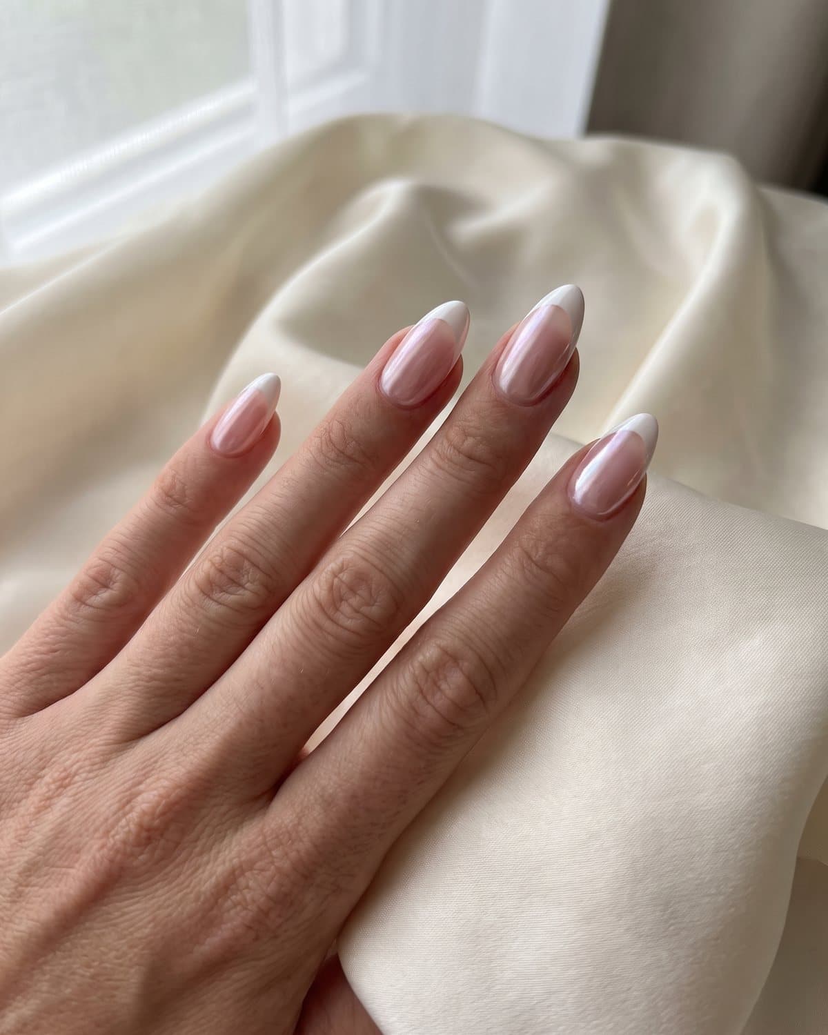 Almond nails with a sheer pink base and white French tips, glazed with pearl chrome, resting on cream silk