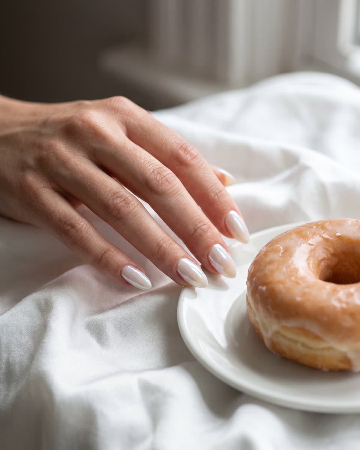 Almond glazed donut nails with a luminous white pearl chrome finish, resting on white silk next to a glazed donut